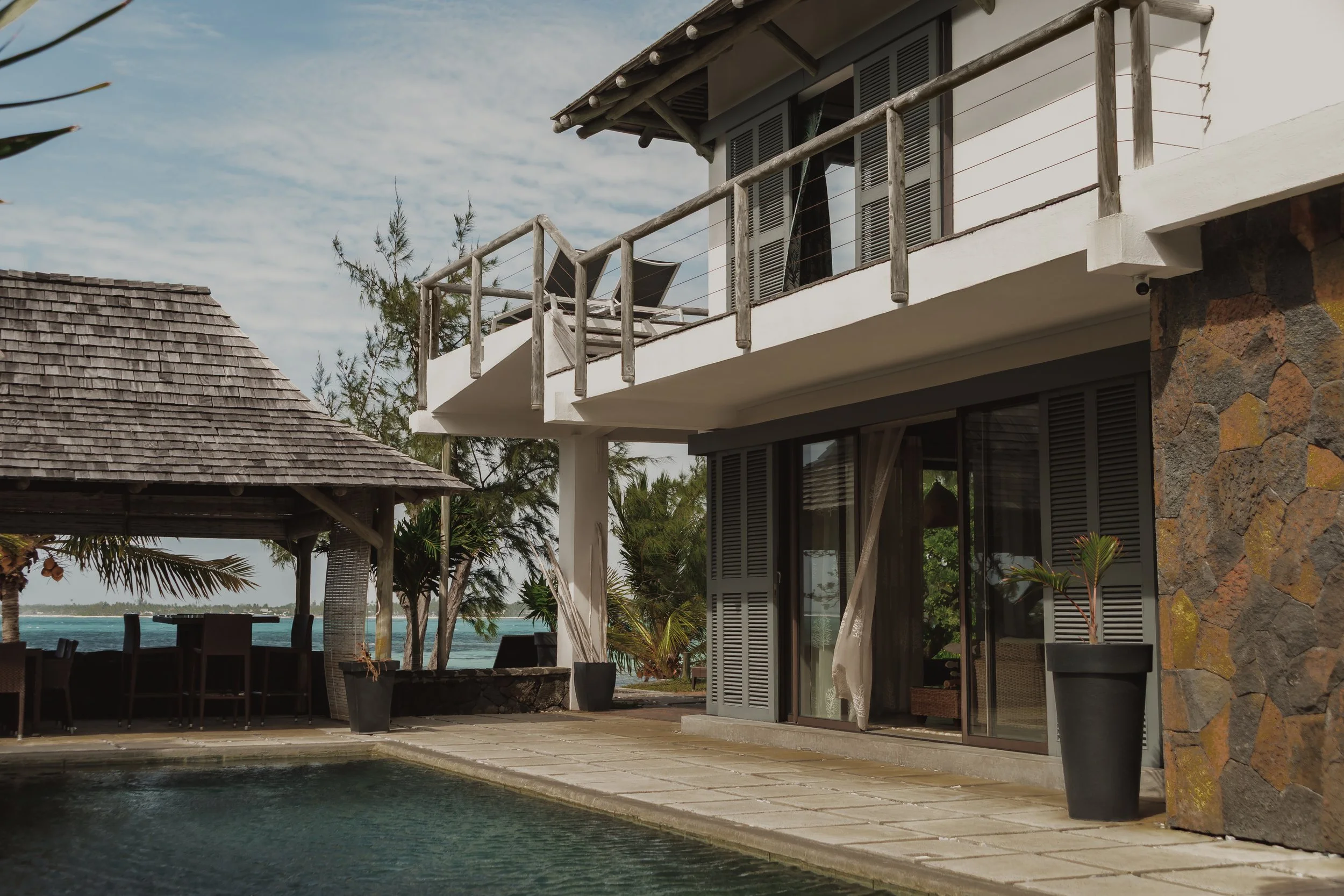 A modern house near the water with a balcony that has lounge chairs, surrounded by tropical plants and trees, with a clear sky in the background.