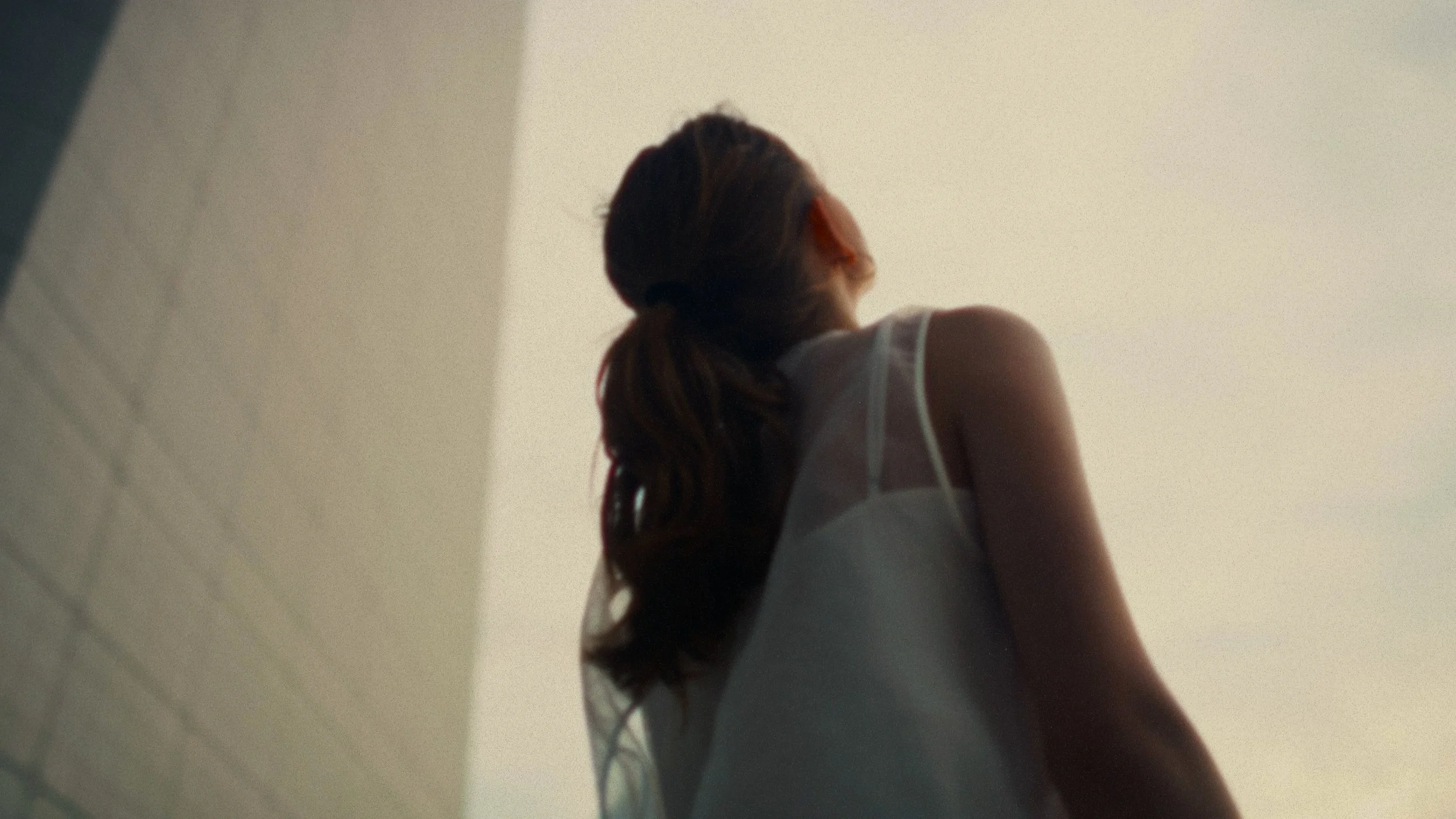 A woman with long hair tied in a ponytail, wearing a white sleeveless top, standing outdoors with a neutral background, viewed from behind and below.