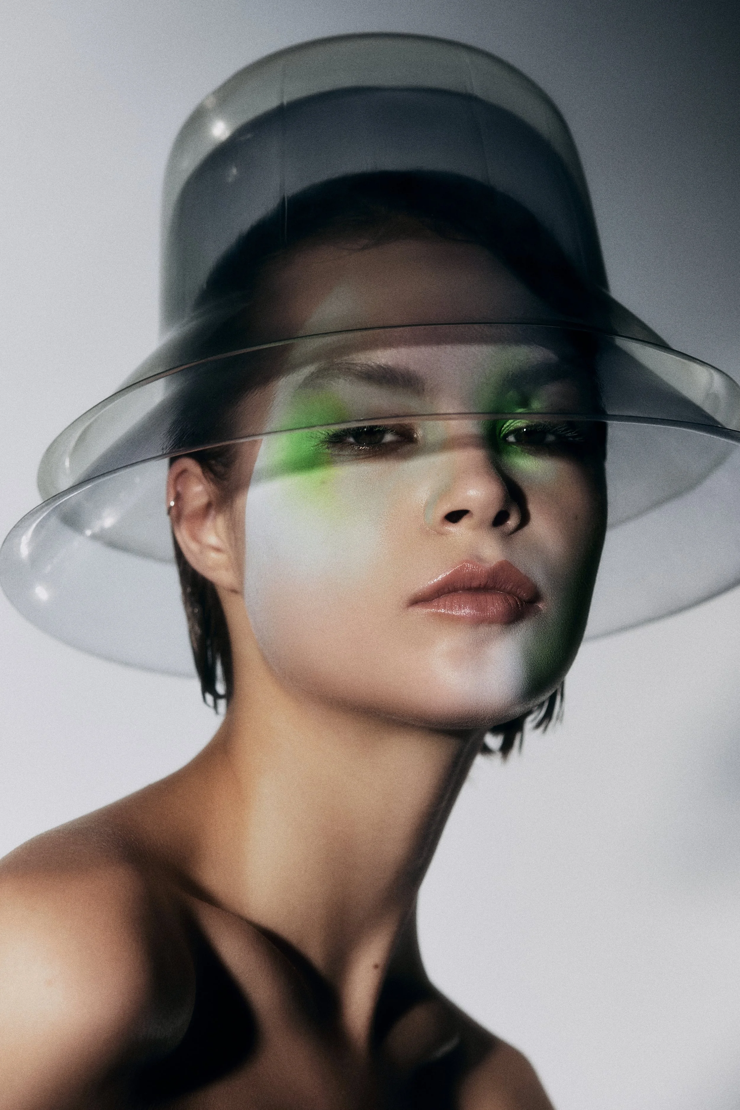 A woman with short dark hair wearing a translucent, layered hat with a futuristic aesthetic, with green lighting reflecting on her face.