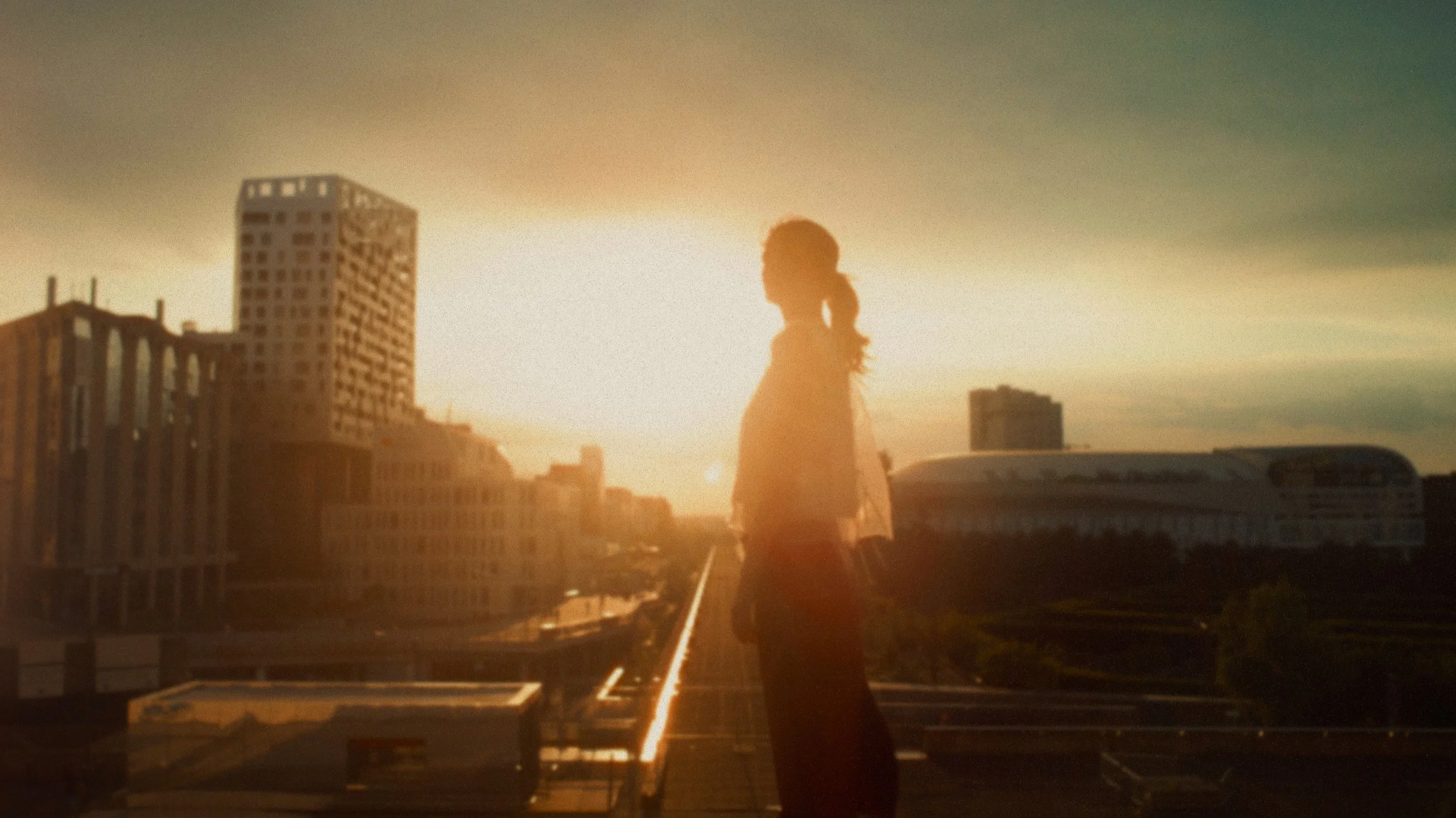 A woman standing on a rooftop during sunset with city buildings in the background.