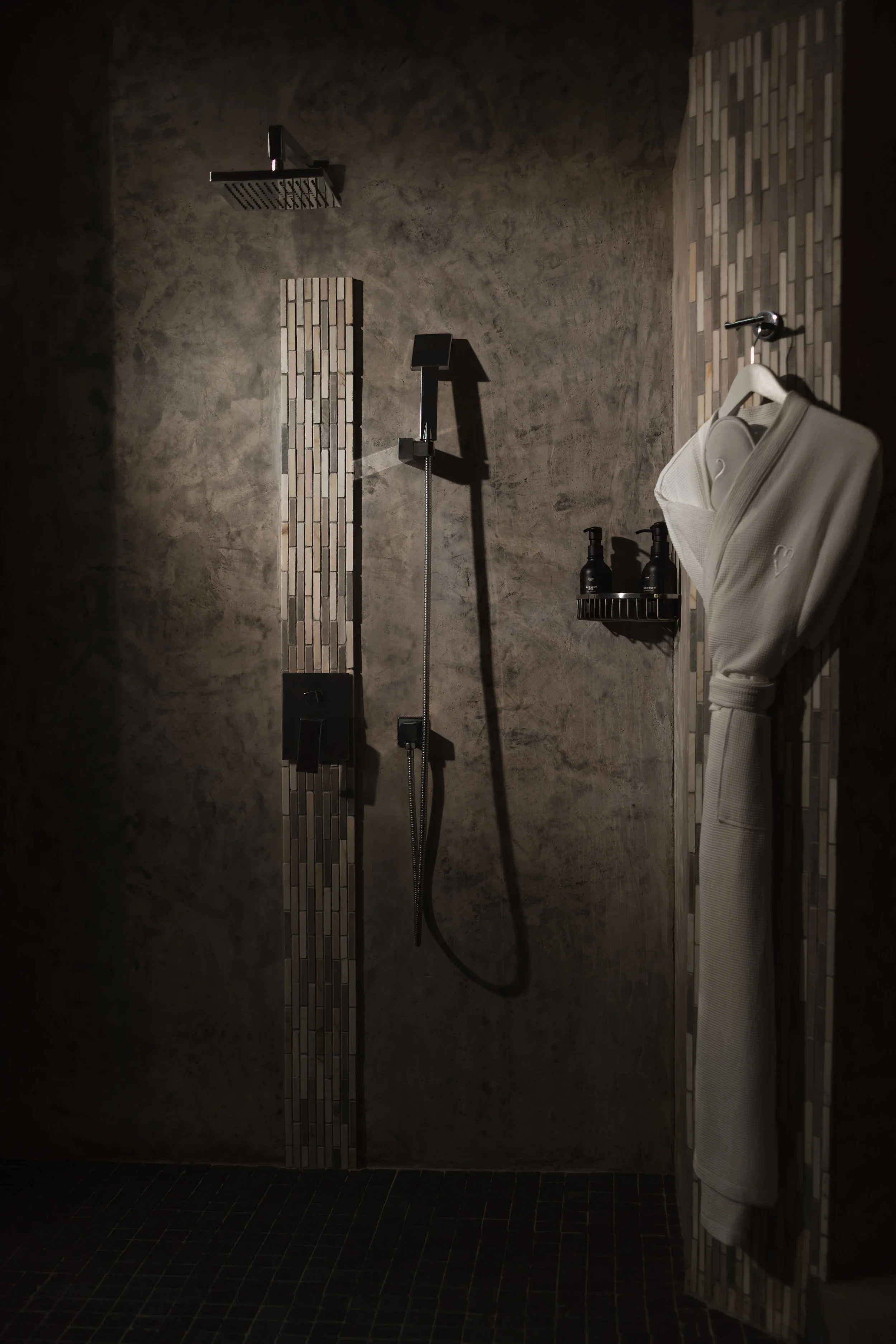A modern shower area with dark tiles on the floor and textured walls. Features include a rainfall showerhead, a handheld shower, a small wall-mounted shelf with two bottles, and a white bathrobe hanging on a hook.