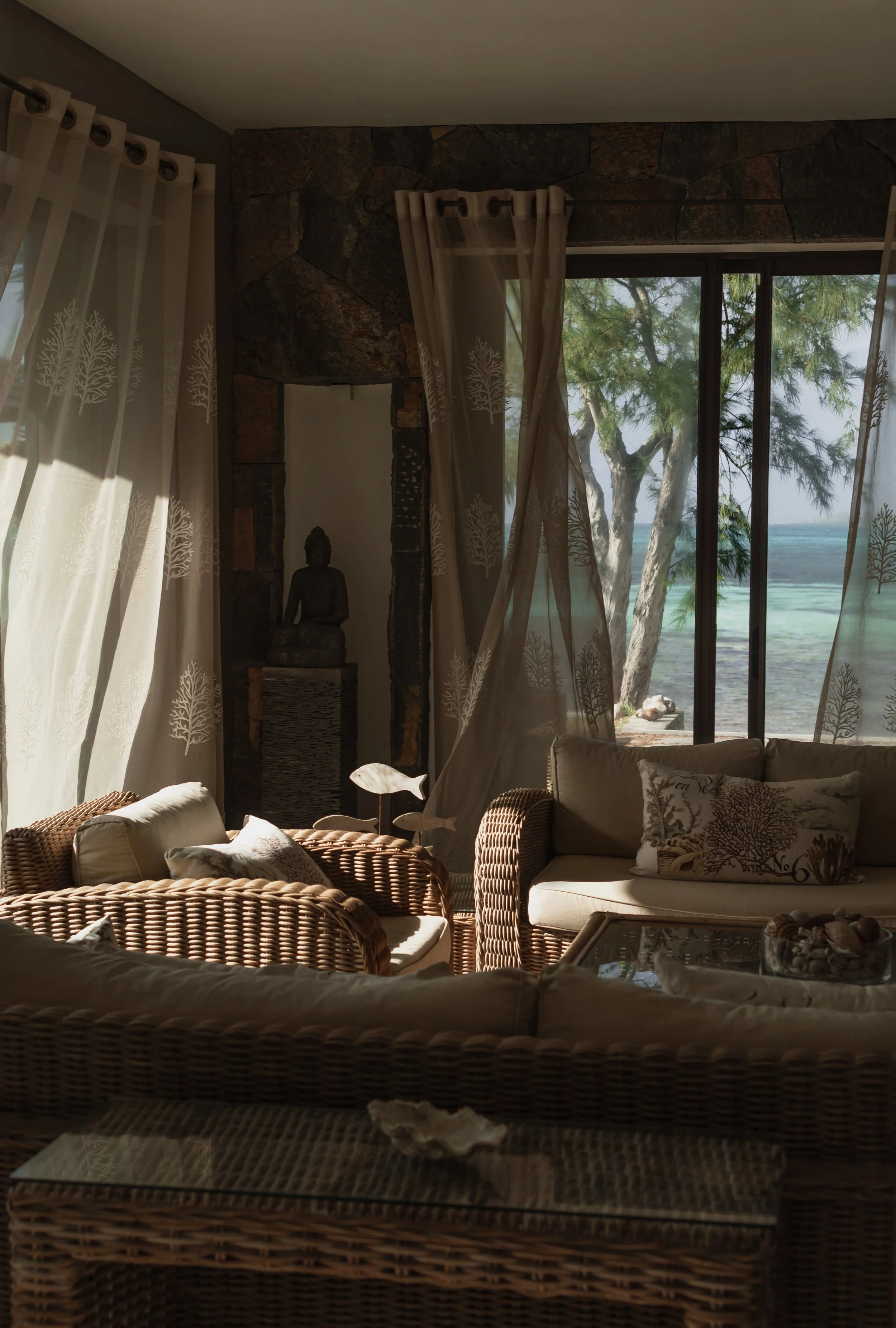 Living room with wicker furniture, white curtains with tree design, a Buddha statue near a stone archway, and large glass door showing trees and water outside.