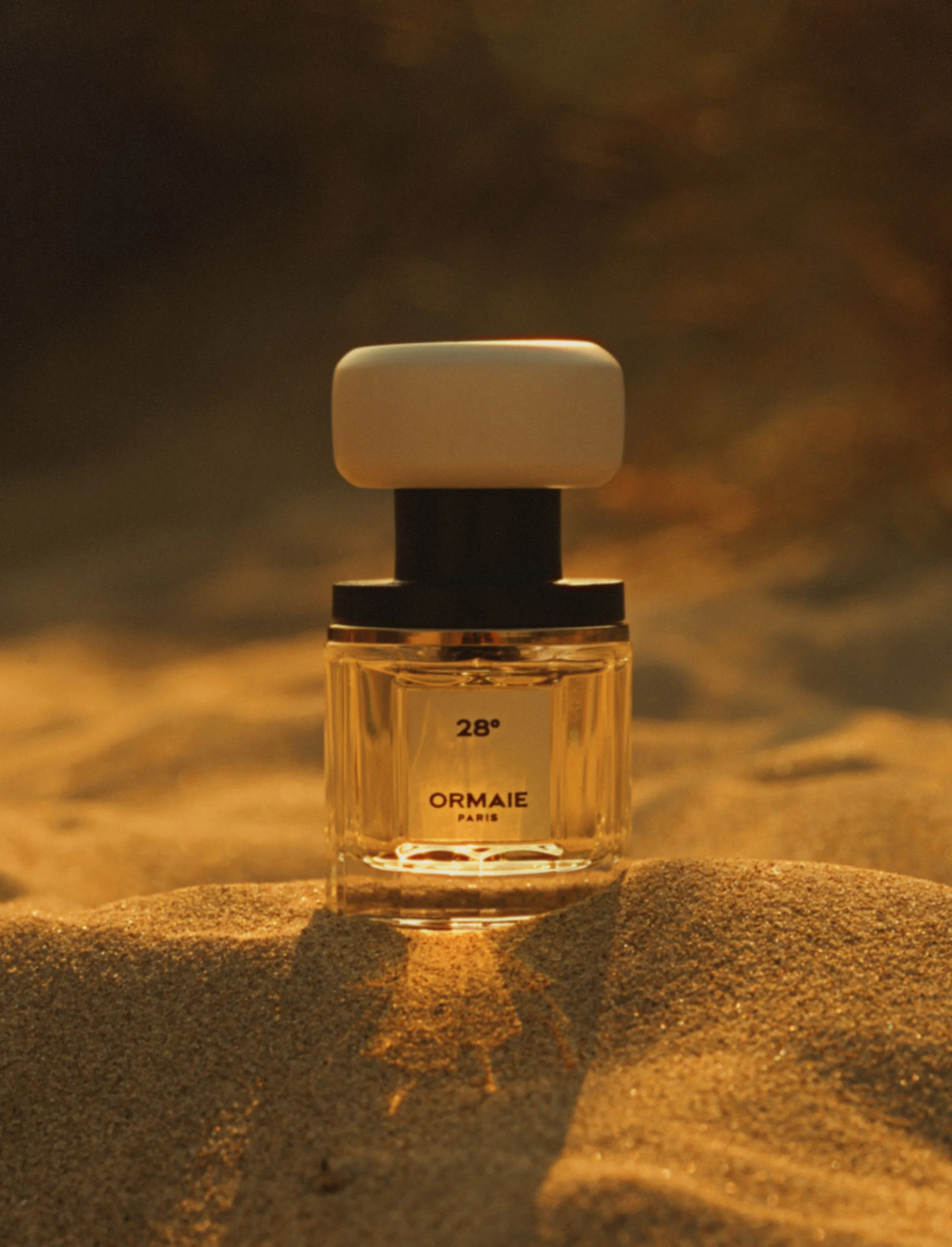 A small perfume bottle with a white cap, placed on sandy ground at sunset or sunrise, with a warm, soft background lighting.