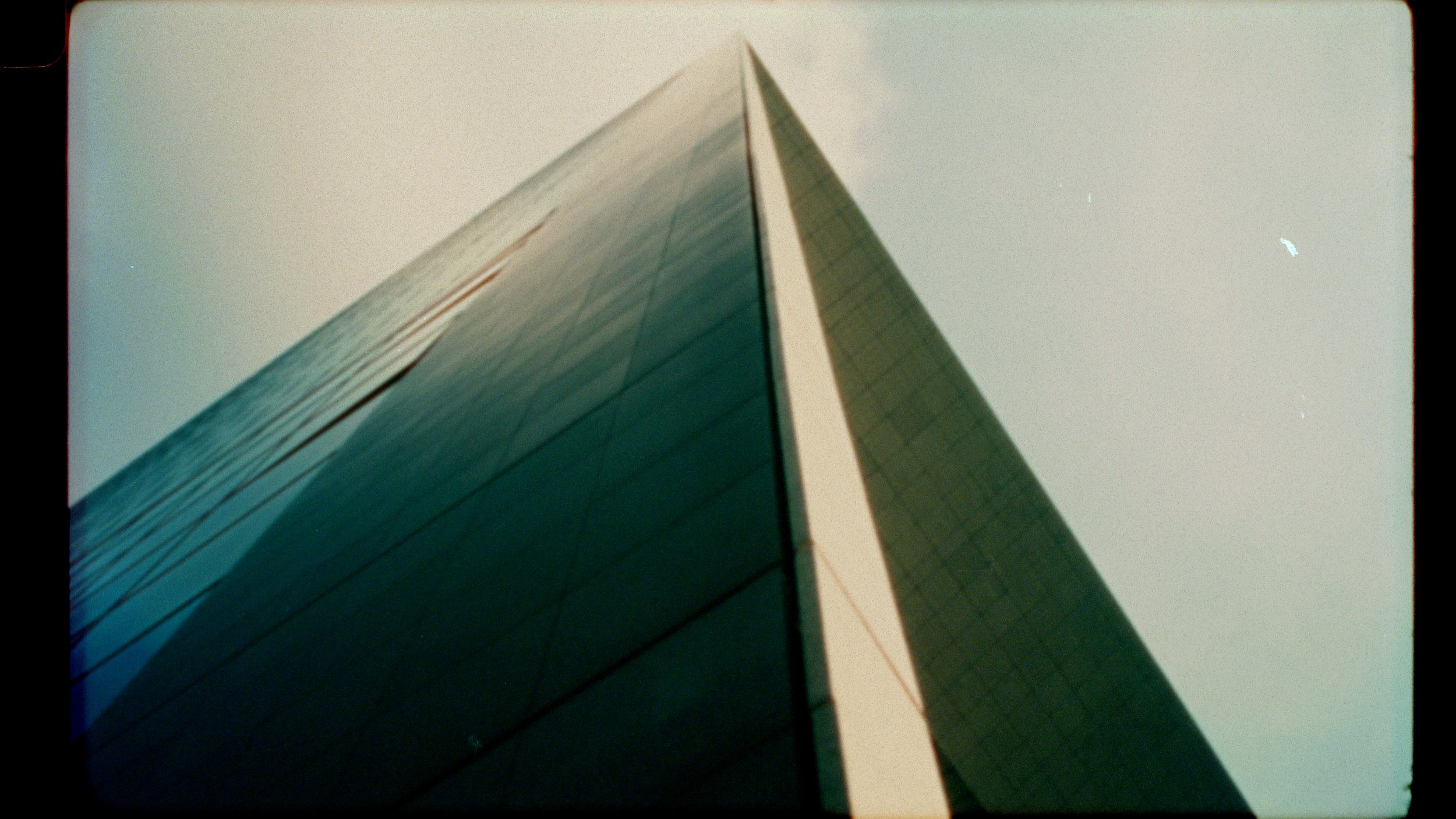 A close-up, upward tilt of a modern high-rise glass building showing its reflective surface and sharp edges, with a pale sky background.