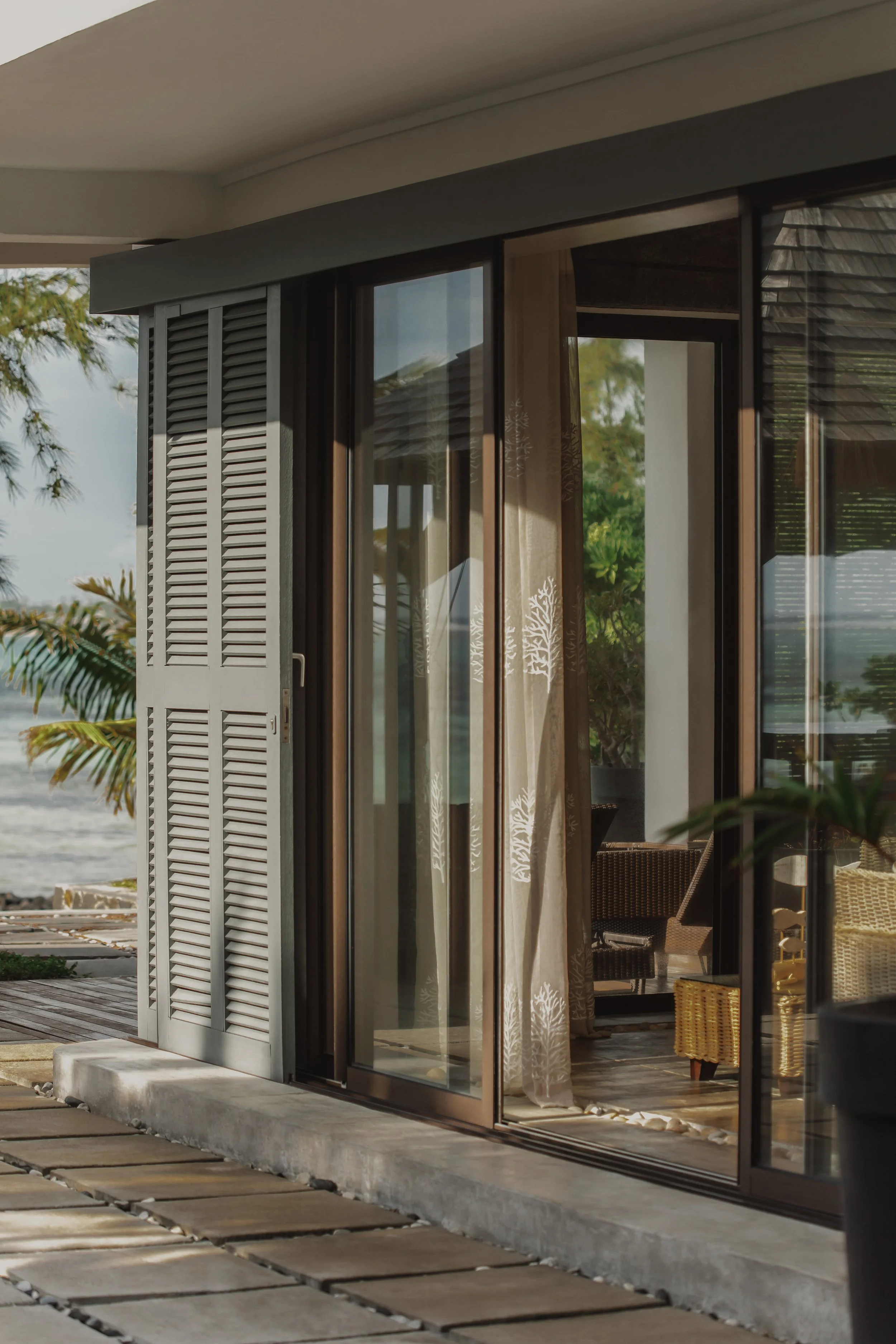 View of a modern house with sliding glass doors, white shutters, and curtains with a nature motif, overlooking a beach and trees outside.