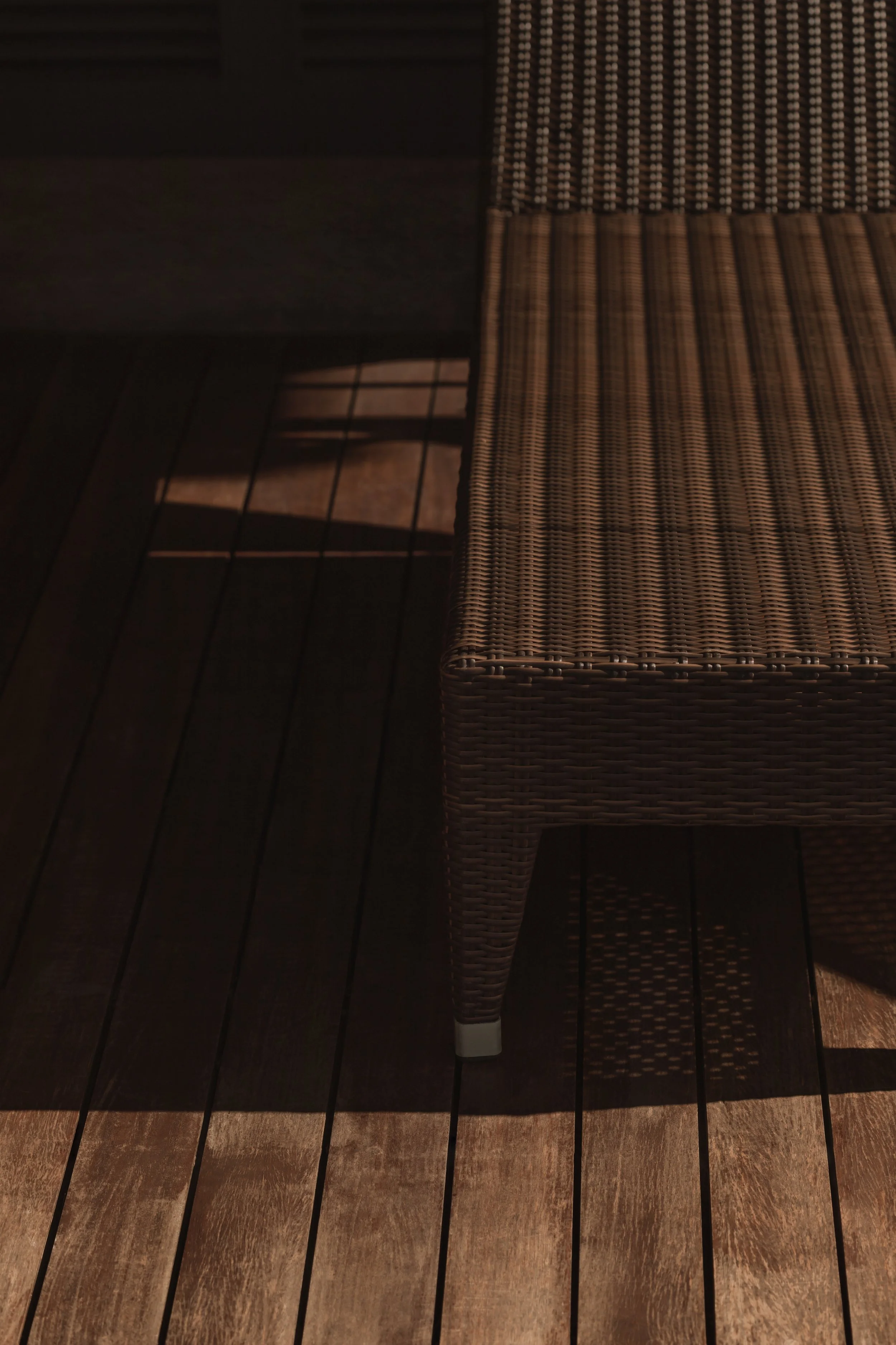 Part of a dark outdoor table and a wicker chair on a wooden deck, sunlight casting shadows.