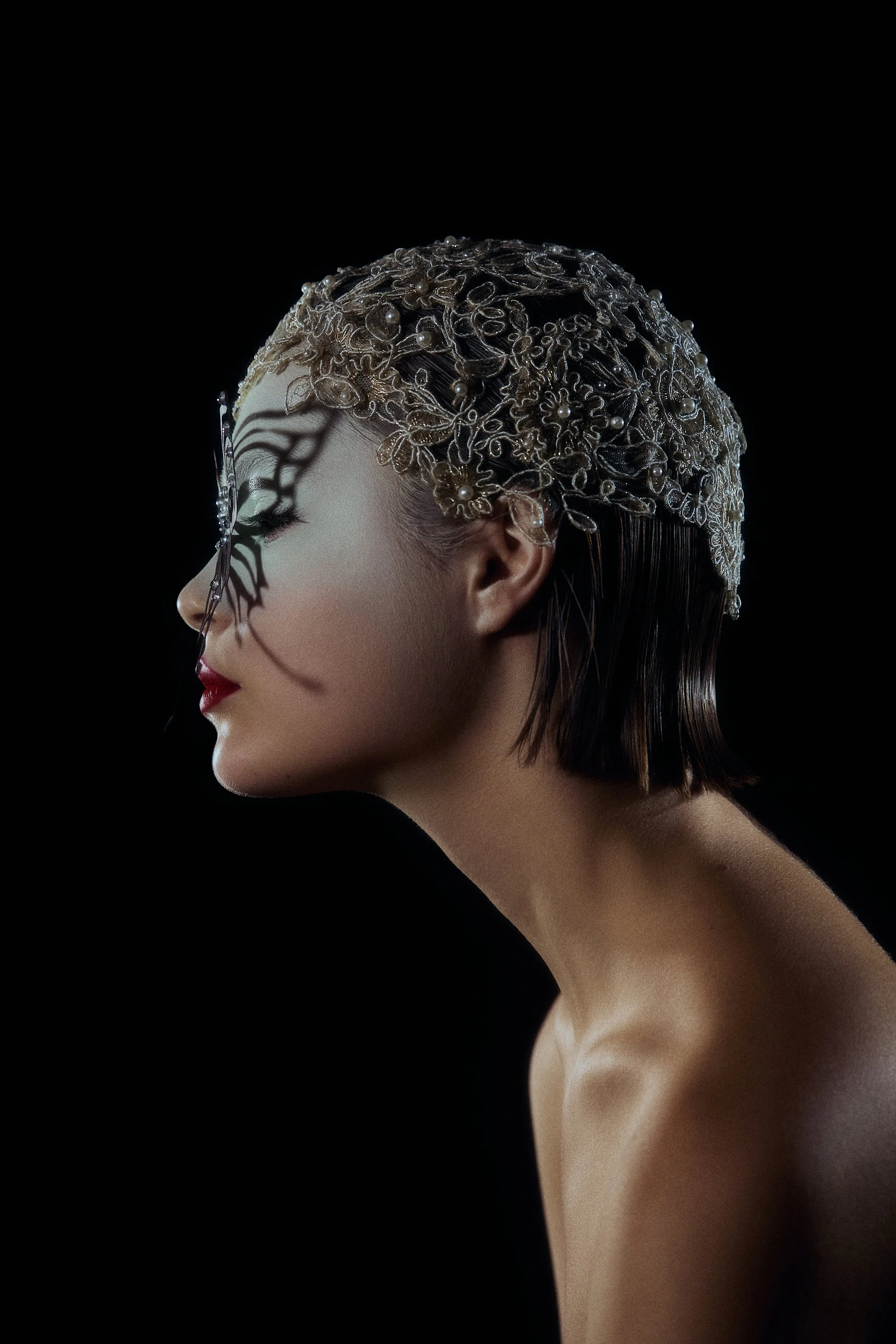 Profile of a woman with short hair wearing an ornate lace headpiece, with a shadow of a butterfly on her face.