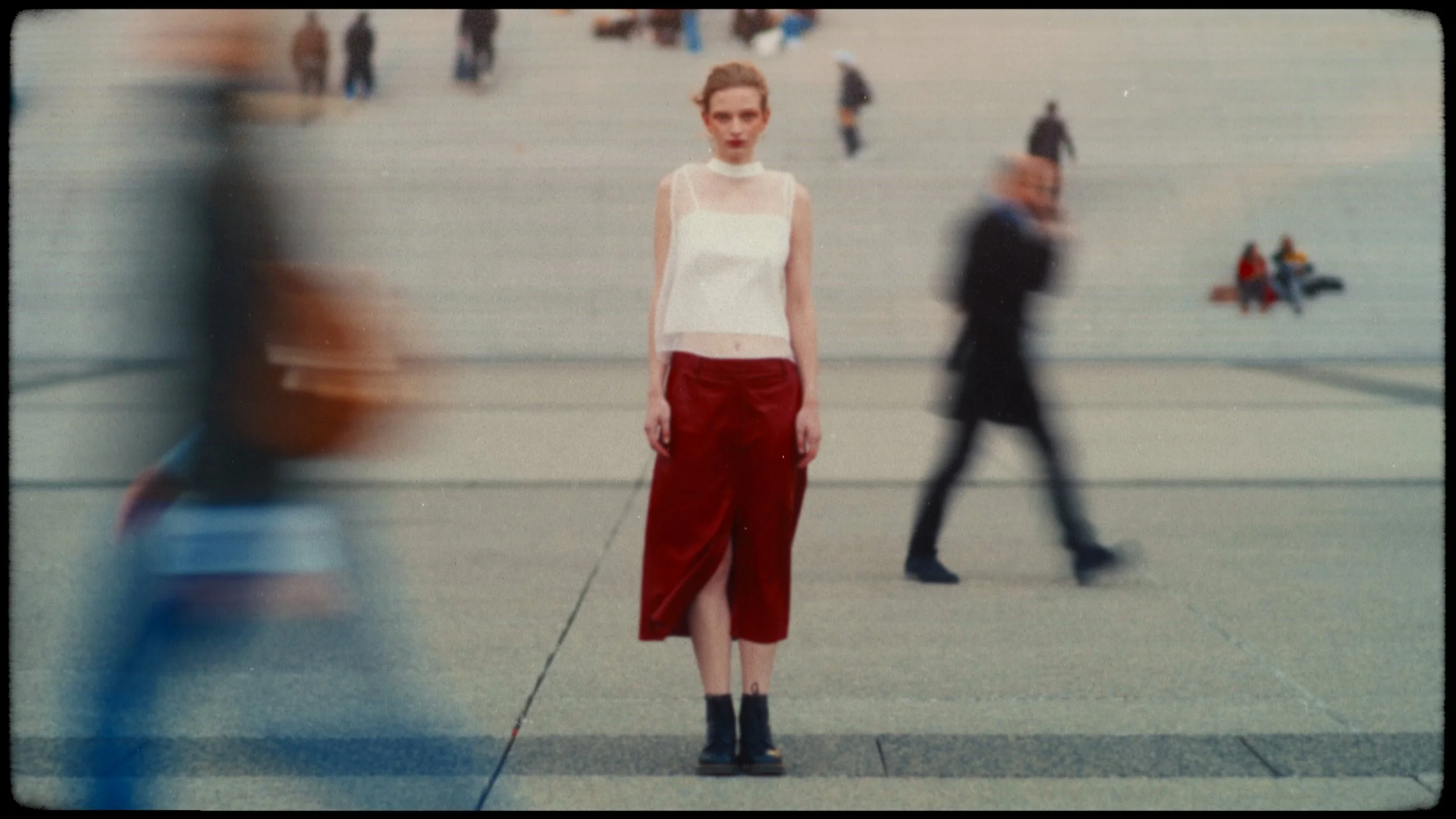 A woman standing still in the middle of a busy city sidewalk, surrounded by blurred pedestrians walking past in different directions.