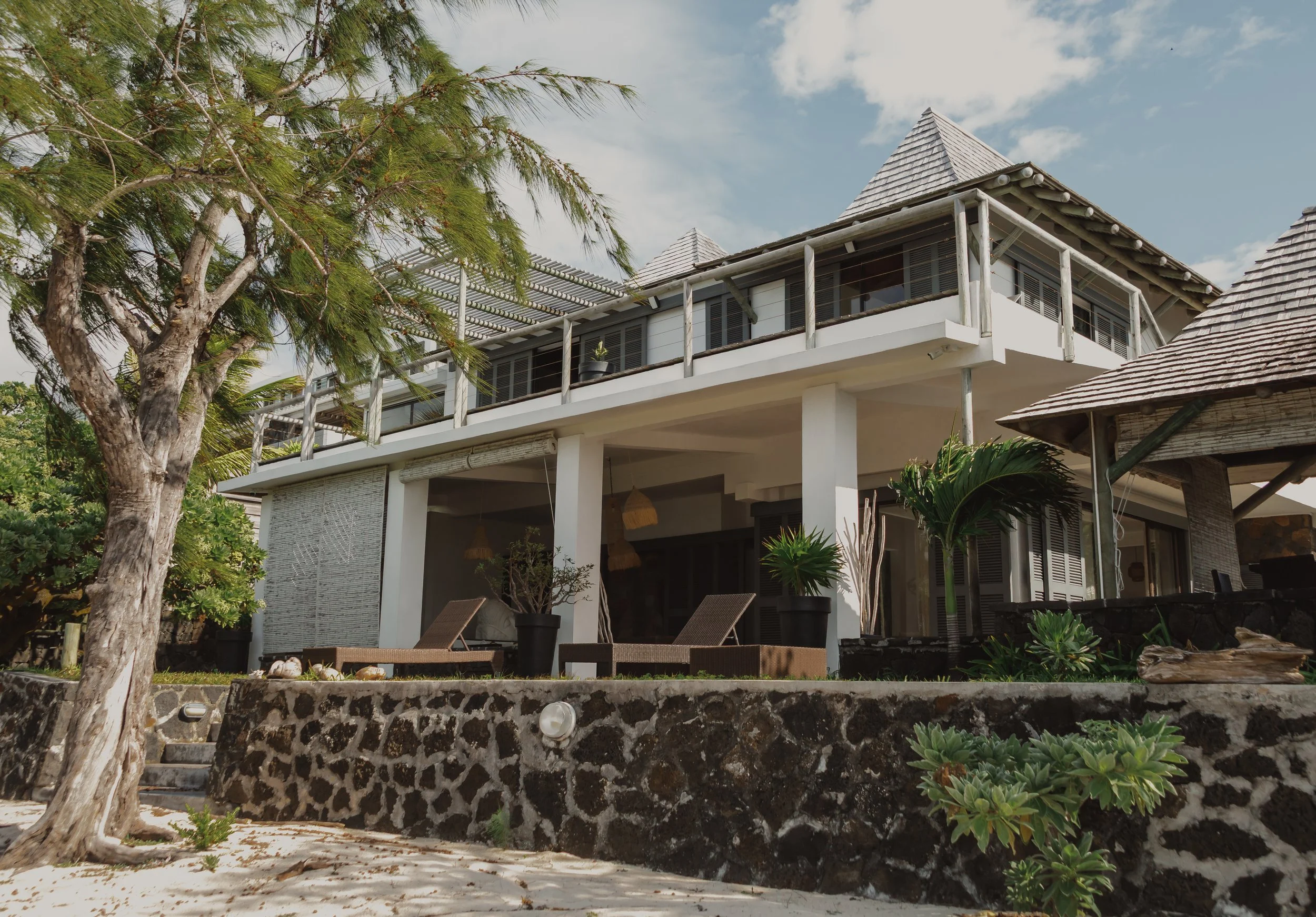 A multi-story house with a balcony, surrounded by palm trees and greenery, with a stone wall and outdoor seating area.