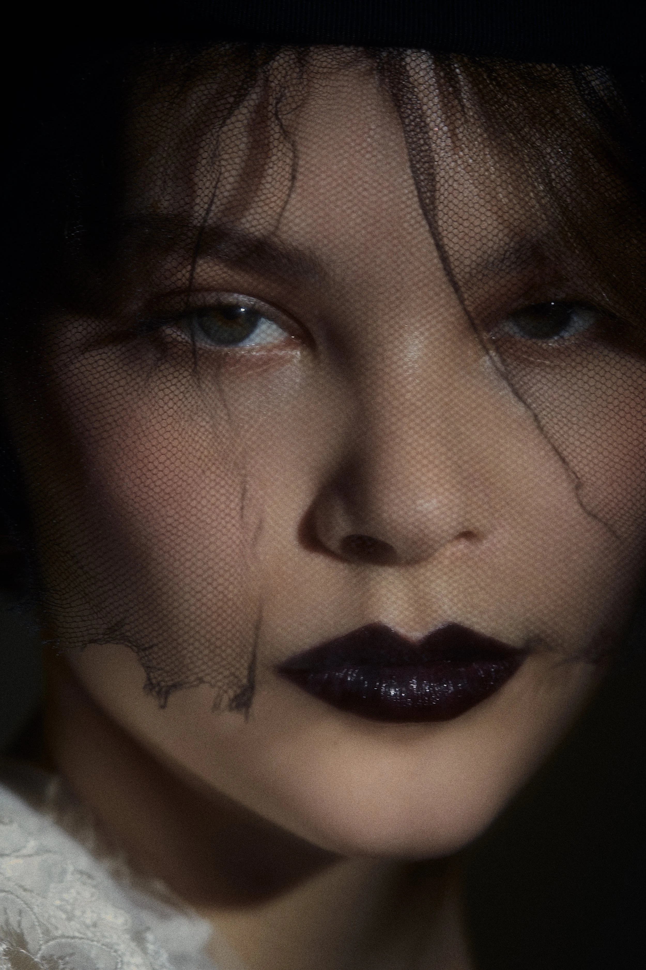 Close-up of a woman with smoky eye makeup and black lipstick, wearing a black veil partially covering her face.