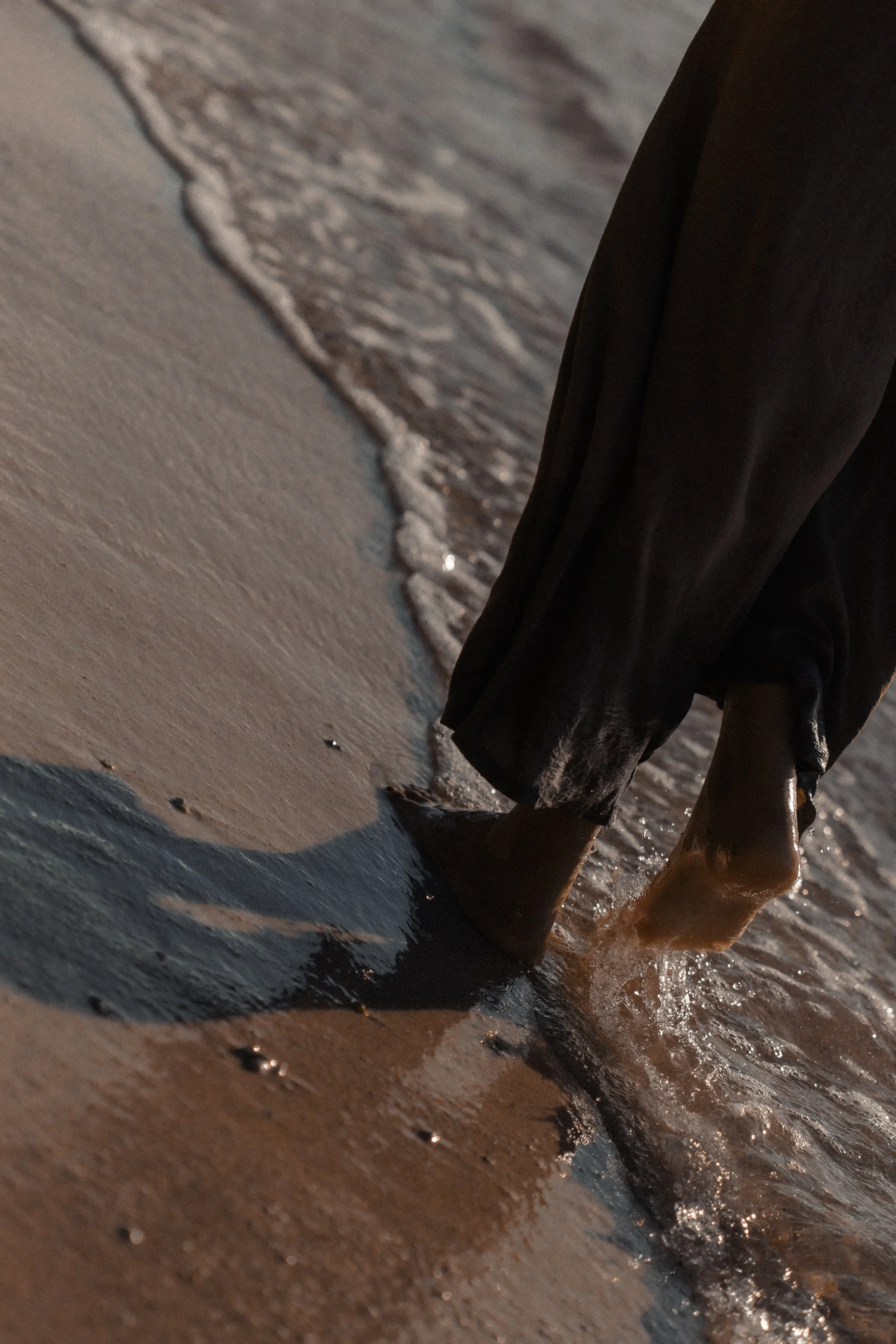 Close-up of bare feet walking along the shoreline with gentle waves washing onto the sandy beach at sunset.