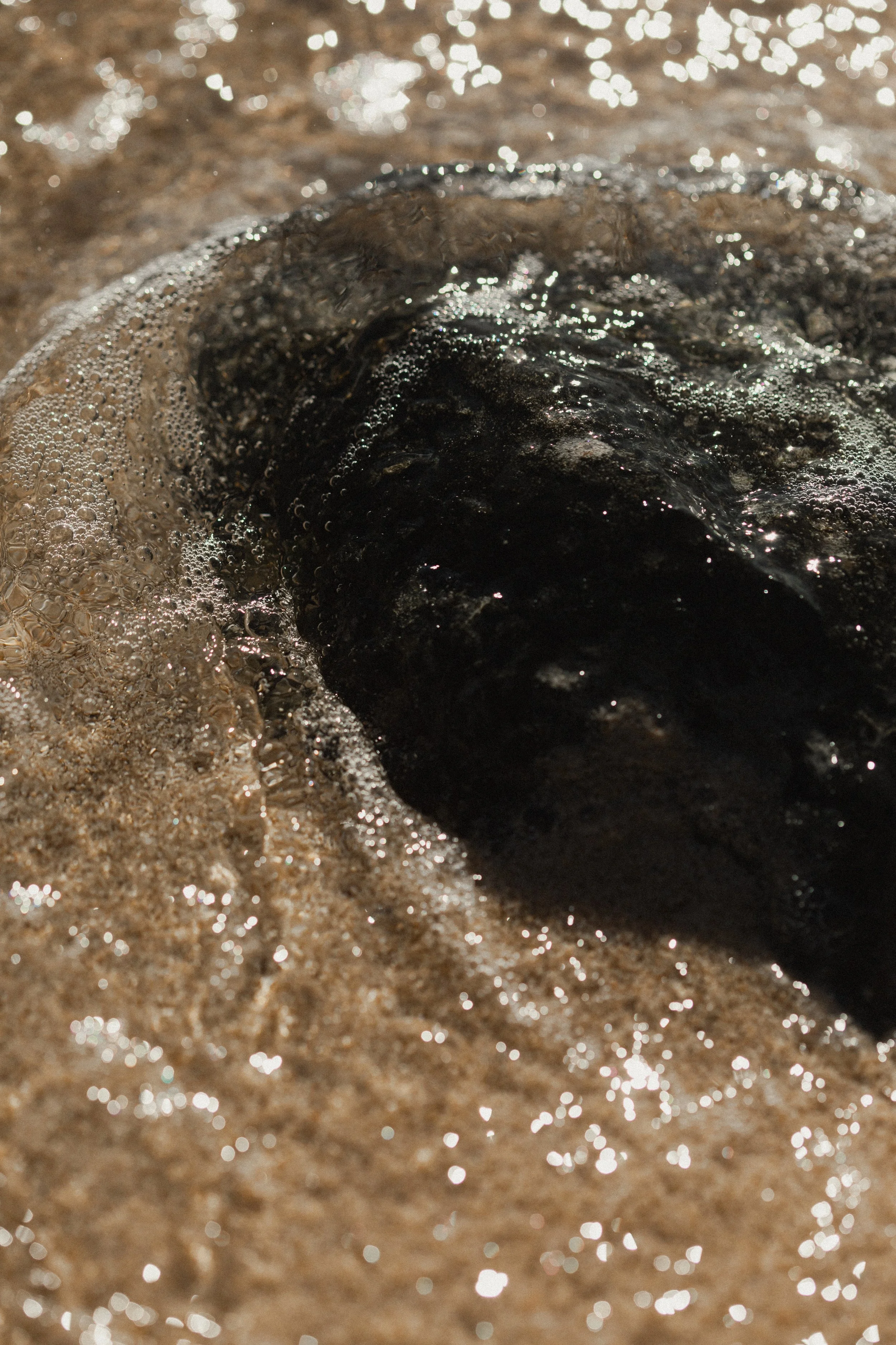 Close-up of a black rock partially submerged in wet sand with small bubbles and shimmering reflections from the sunlight.