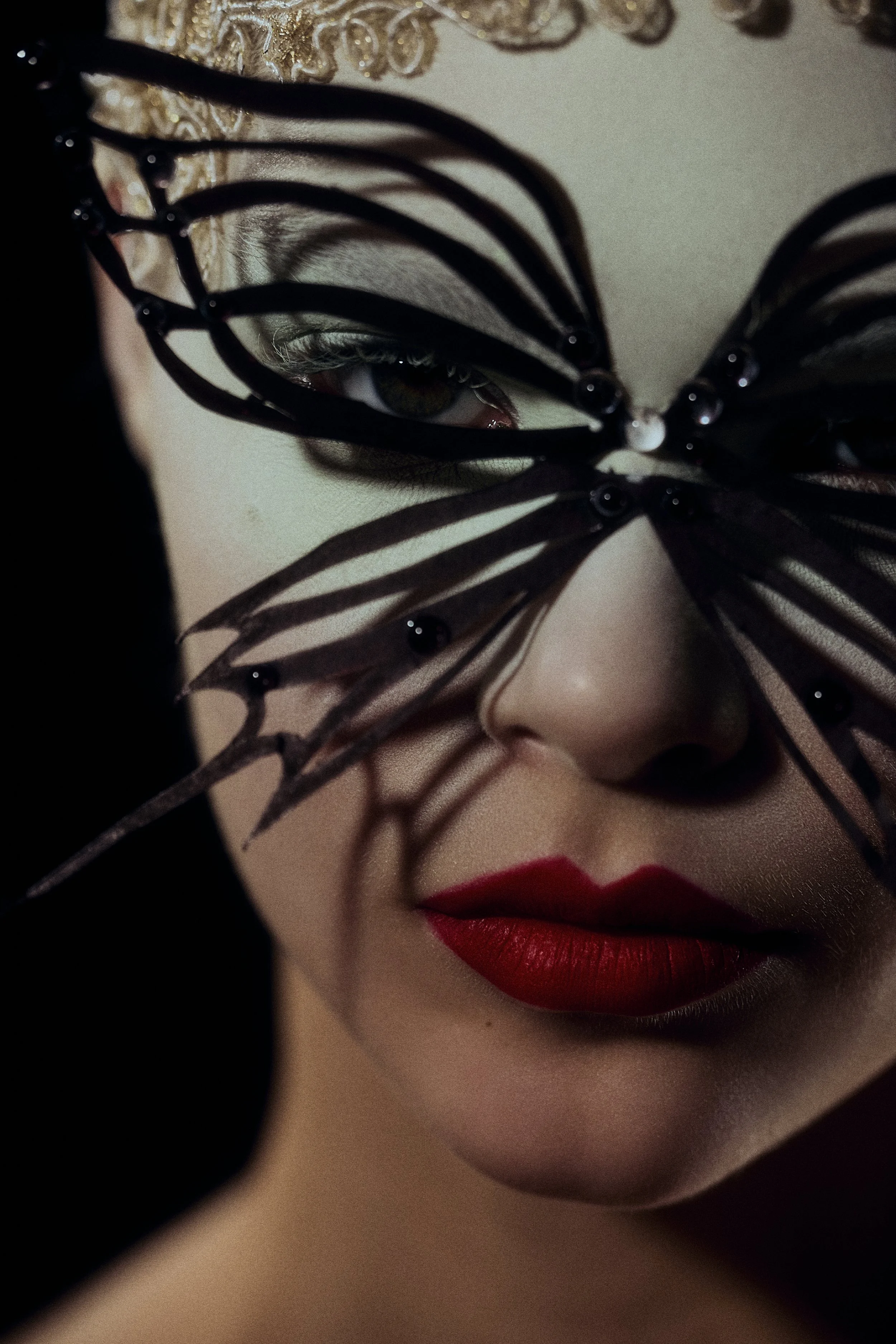 Close-up of a woman's face with red lipstick, partially covered by a black lace mask with beads, and gold embroidery on her forehead.