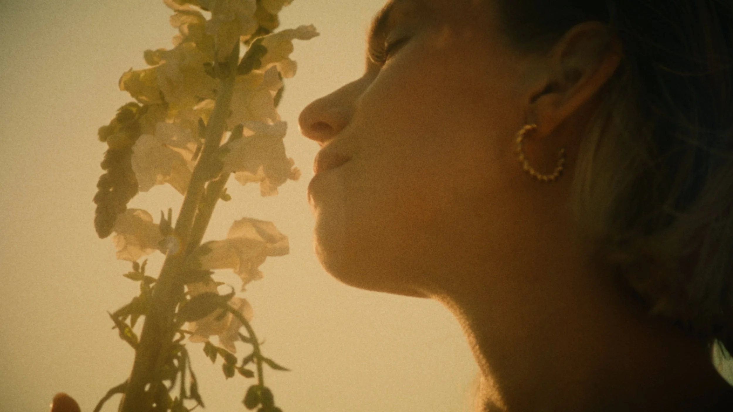 A woman with earrings is posing near a flowering tree with pale pink or white blossoms, with warm lighting and a beige background.