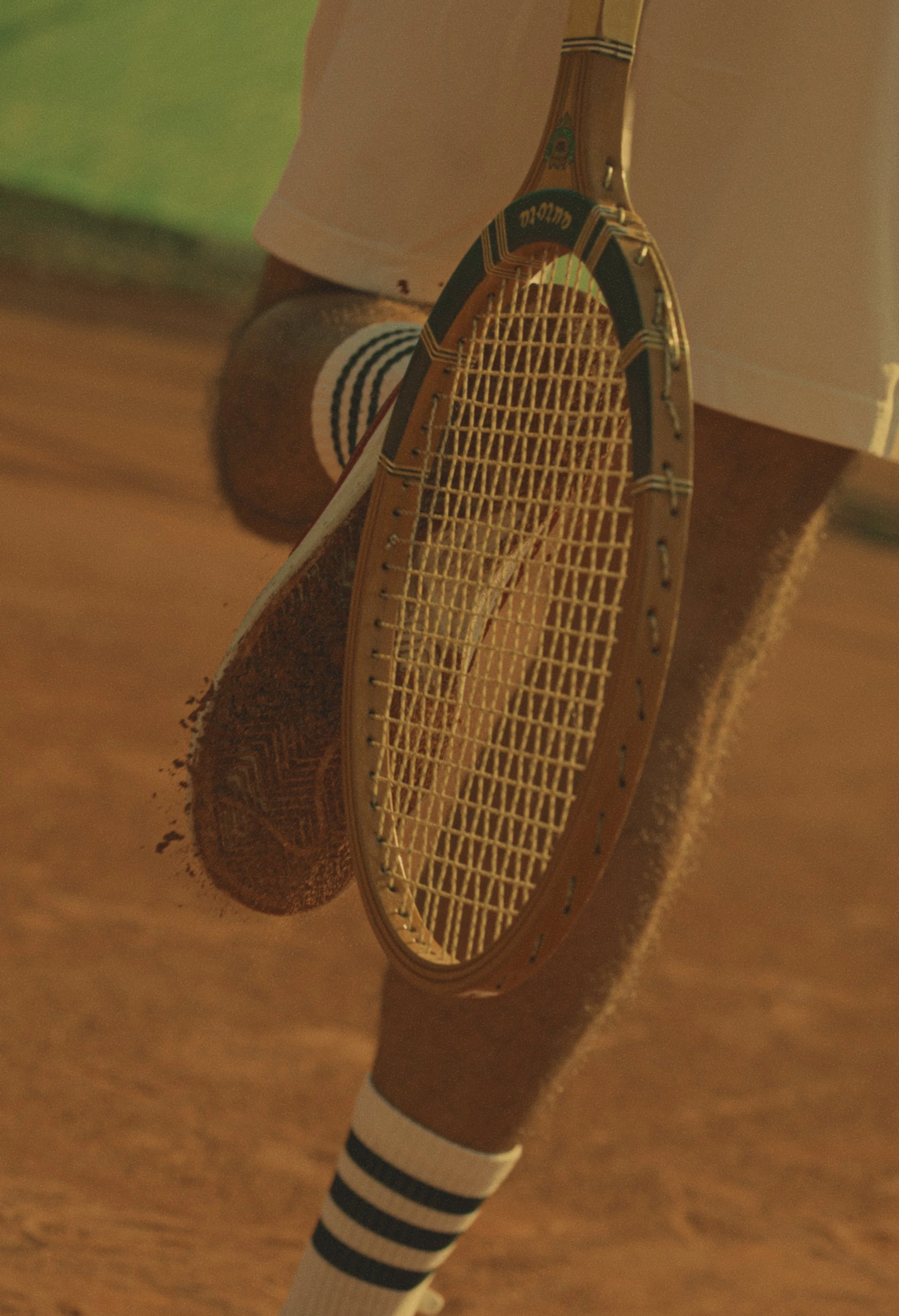 A tennis racket leaning against a person's legs, who is wearing white shorts and black and white striped socks; a tennis ball and tennis shoes are visible on a wooden court.