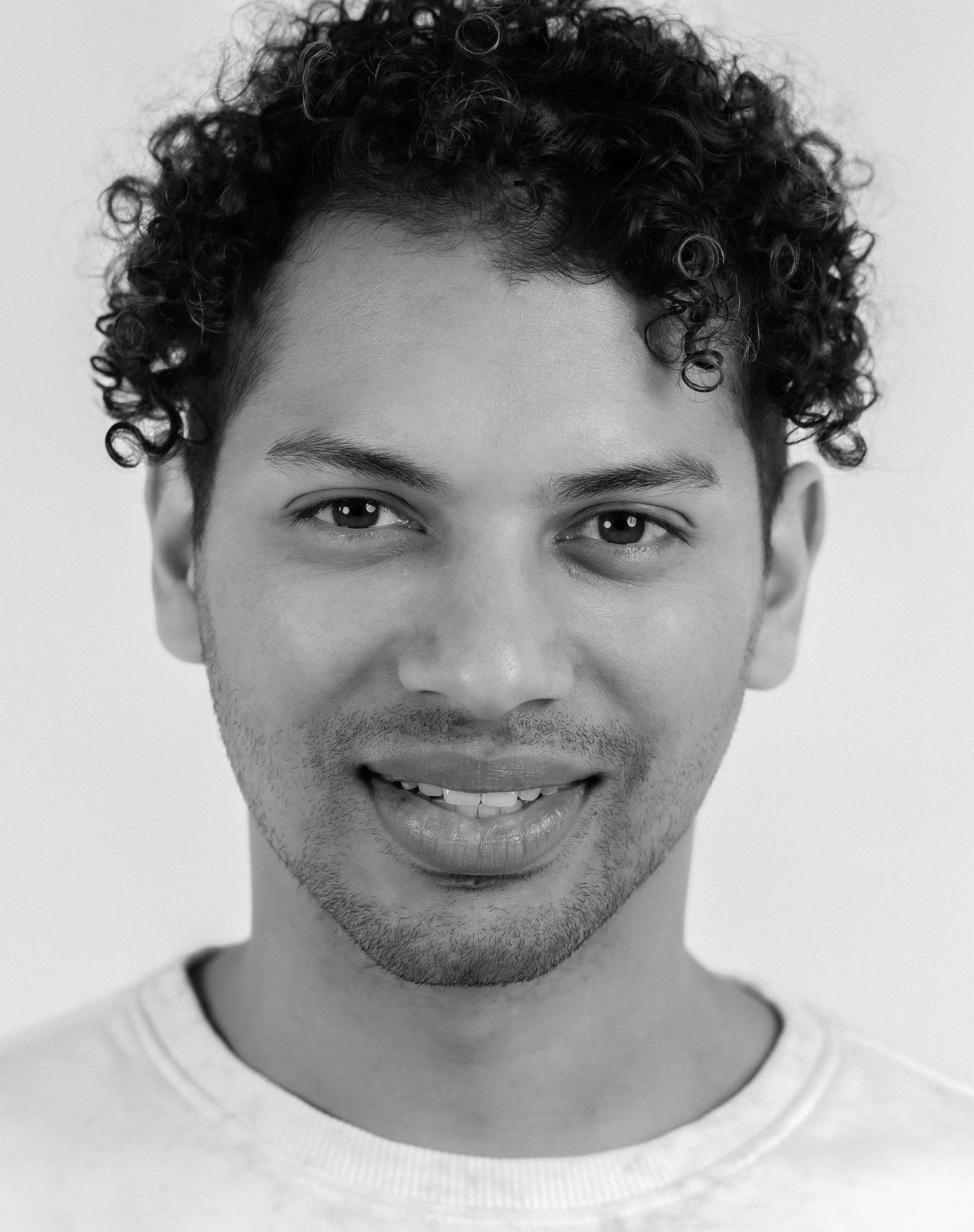 A smiling young man with curly hair and a light beard, wearing a light-colored T-shirt, posing against a plain background.