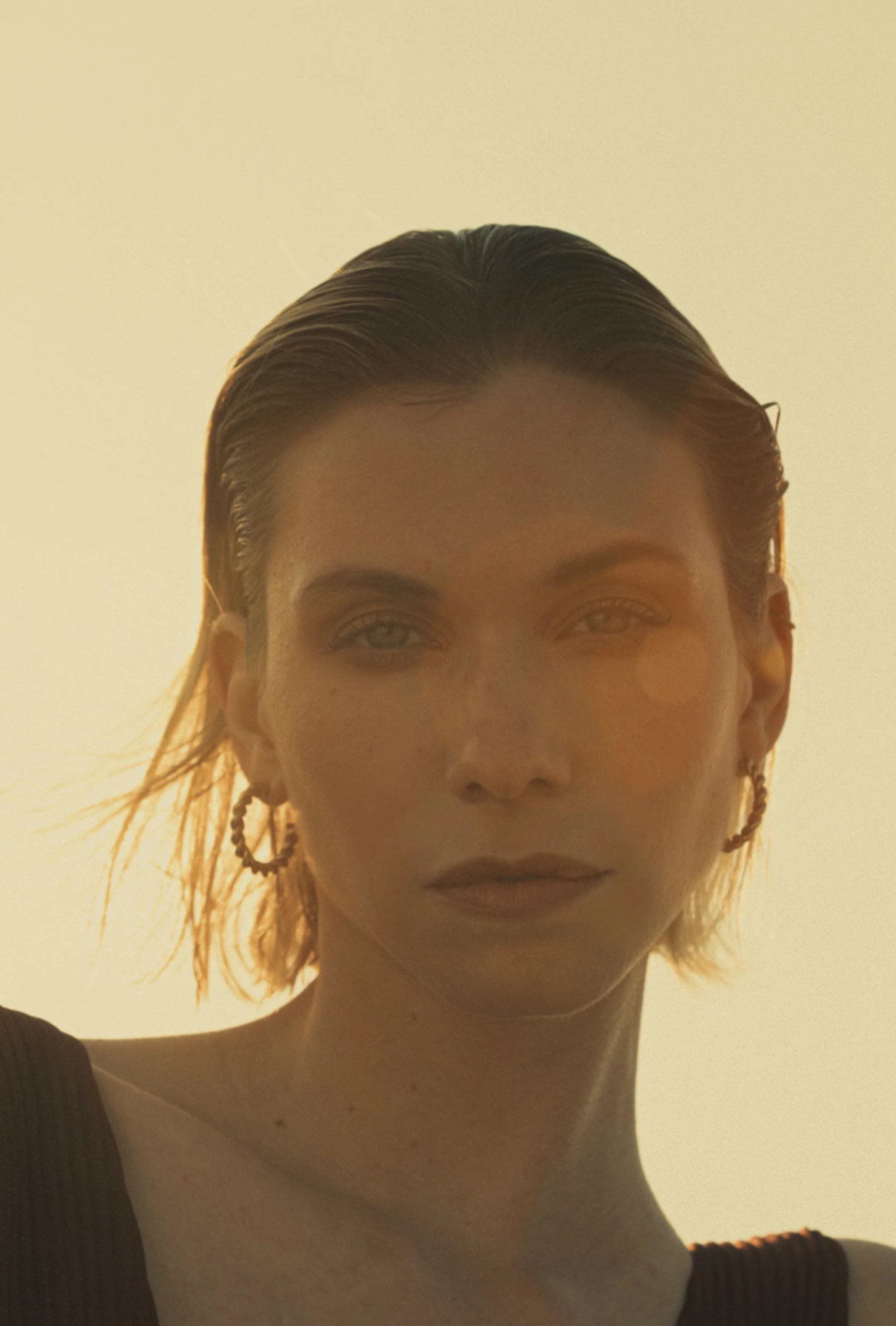 Close-up of a person with wet, slicked-back hair, lightly tanned skin, and wearing a sleeveless black top. The person has light makeup, hoop earrings, and faint freckles.