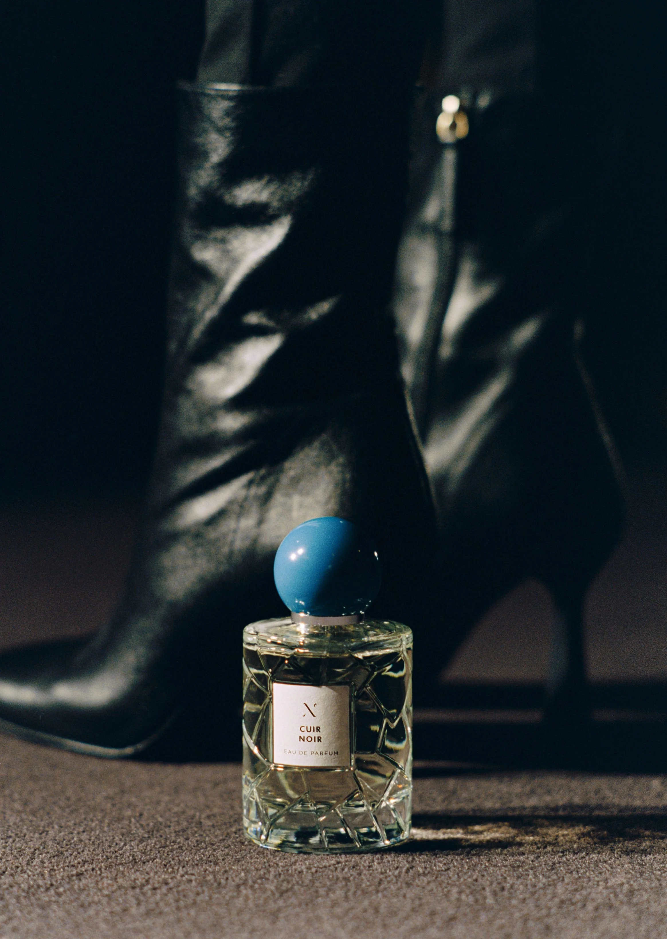 A perfume bottle labeled 'Cuir Noir' with a blue spherical cap, placed on a textured surface with a black fabric backdrop.