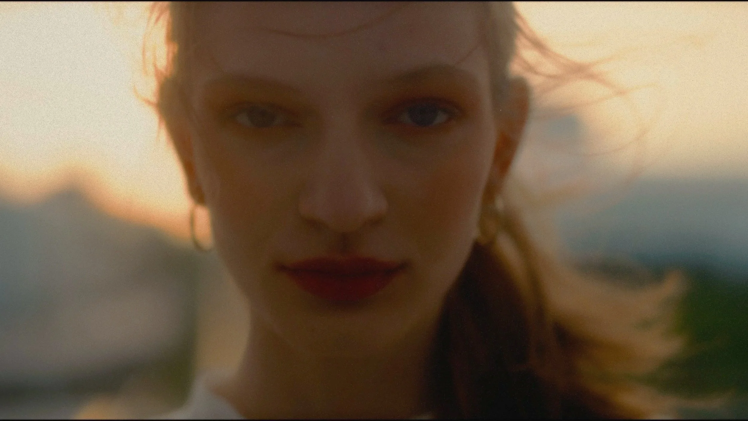 A close-up of a young woman with red hair and blue eyes outdoors during sunset, with wind blowing strands of her hair.