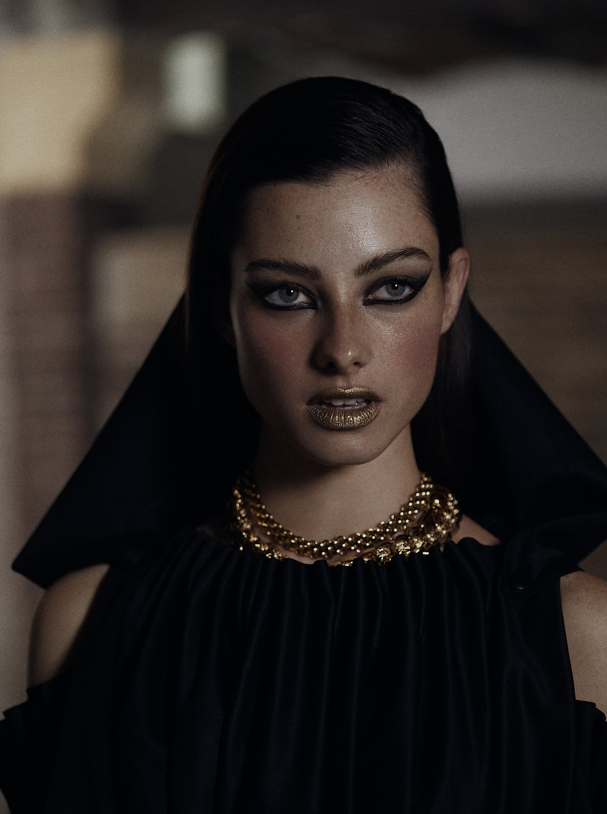 A woman with dark hair, bold makeup, and gold lipstick, wearing a black ruffled dress with a large collar and layered gold necklaces, looking directly at the camera.