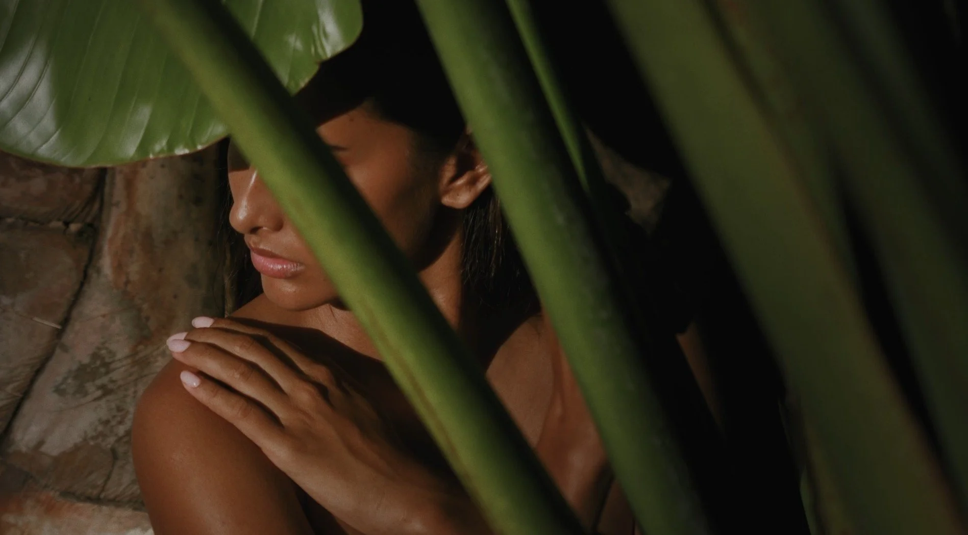 Woman with medium skin tone, dark hair, and closed eyes, partially obscured by large green tropical leaves, against a brick wall background.