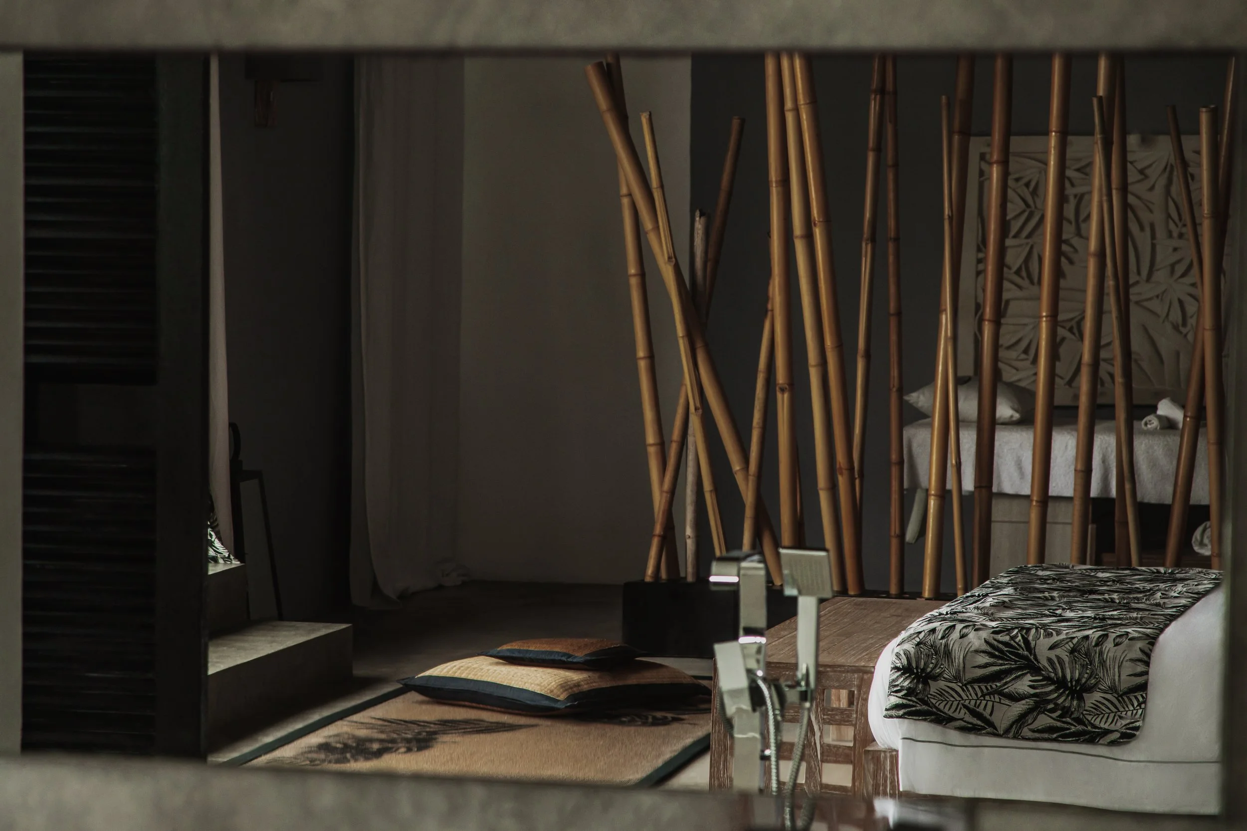 A bedroom view through a mirror showing a bed with black and white leafy patterned pillows, a white bedspread, a small table with an item on it, and a wall with decorative wood or bamboo sticks.