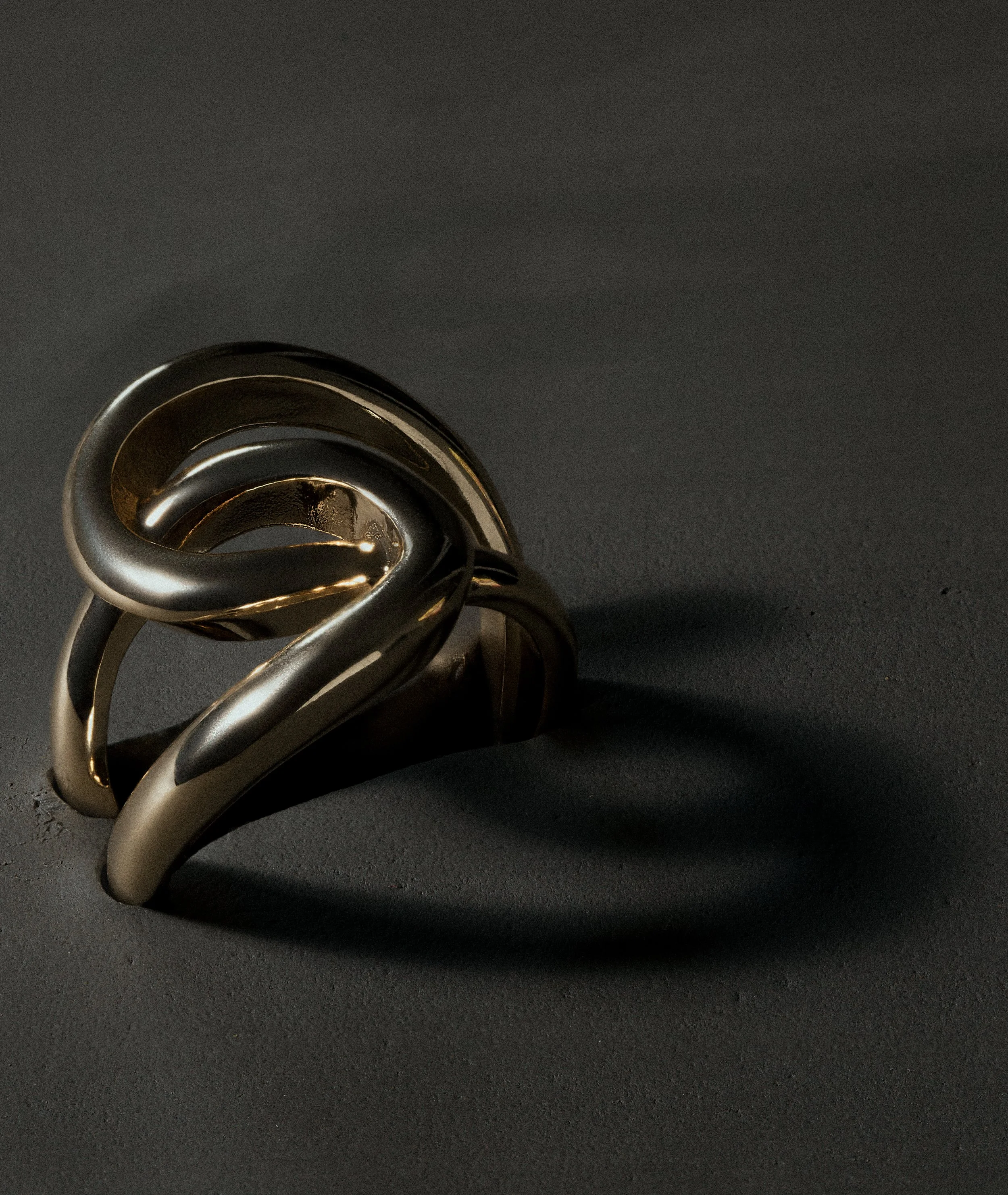 A shiny metallic ring with a modern, abstract design placed on a dark surface, casting a shadow.