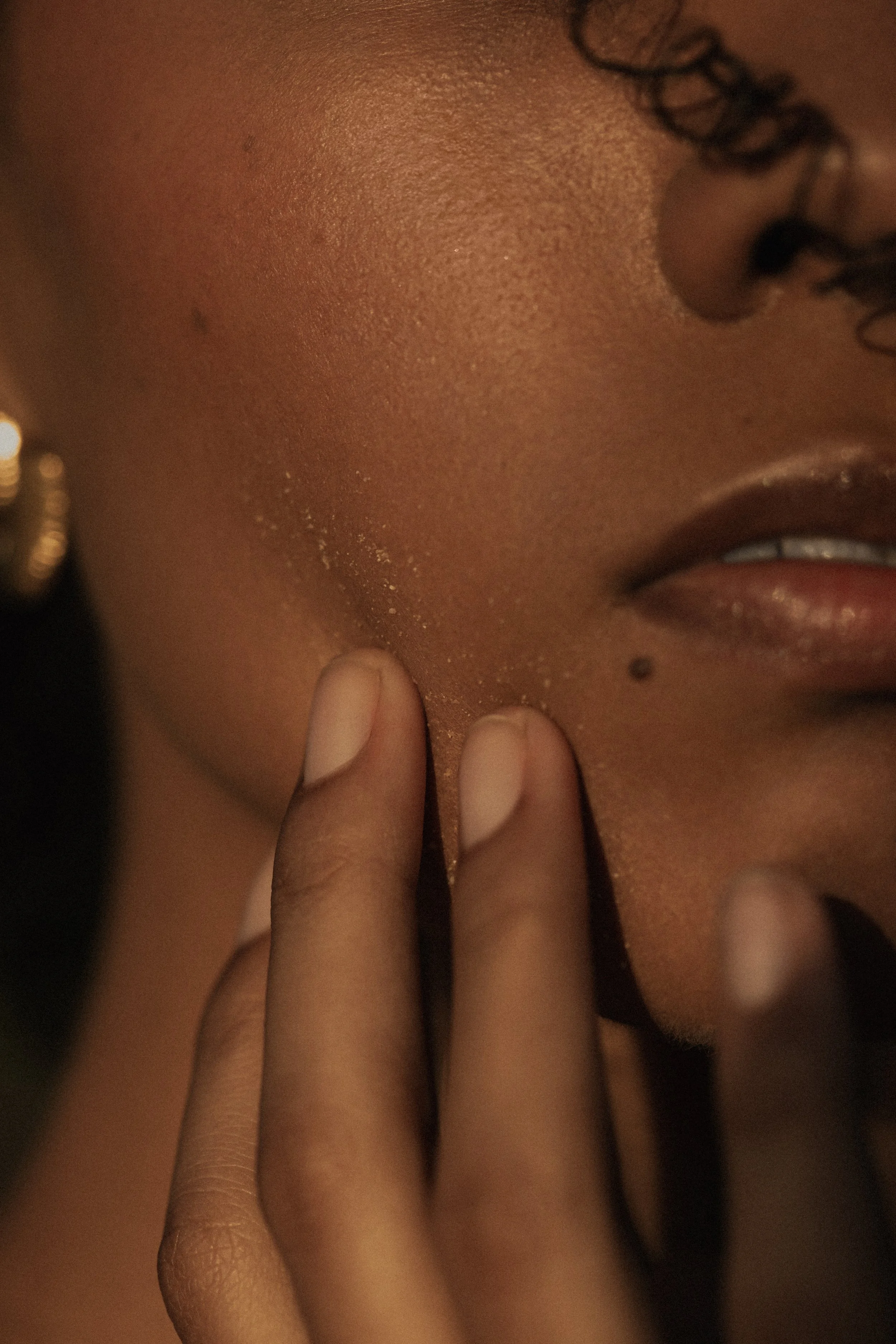 Close-up of a person's face focusing on their cheek, part of their nose, lips, and a hand touching their chin. The person has dark skin, curly hair, and is wearing a small earring.