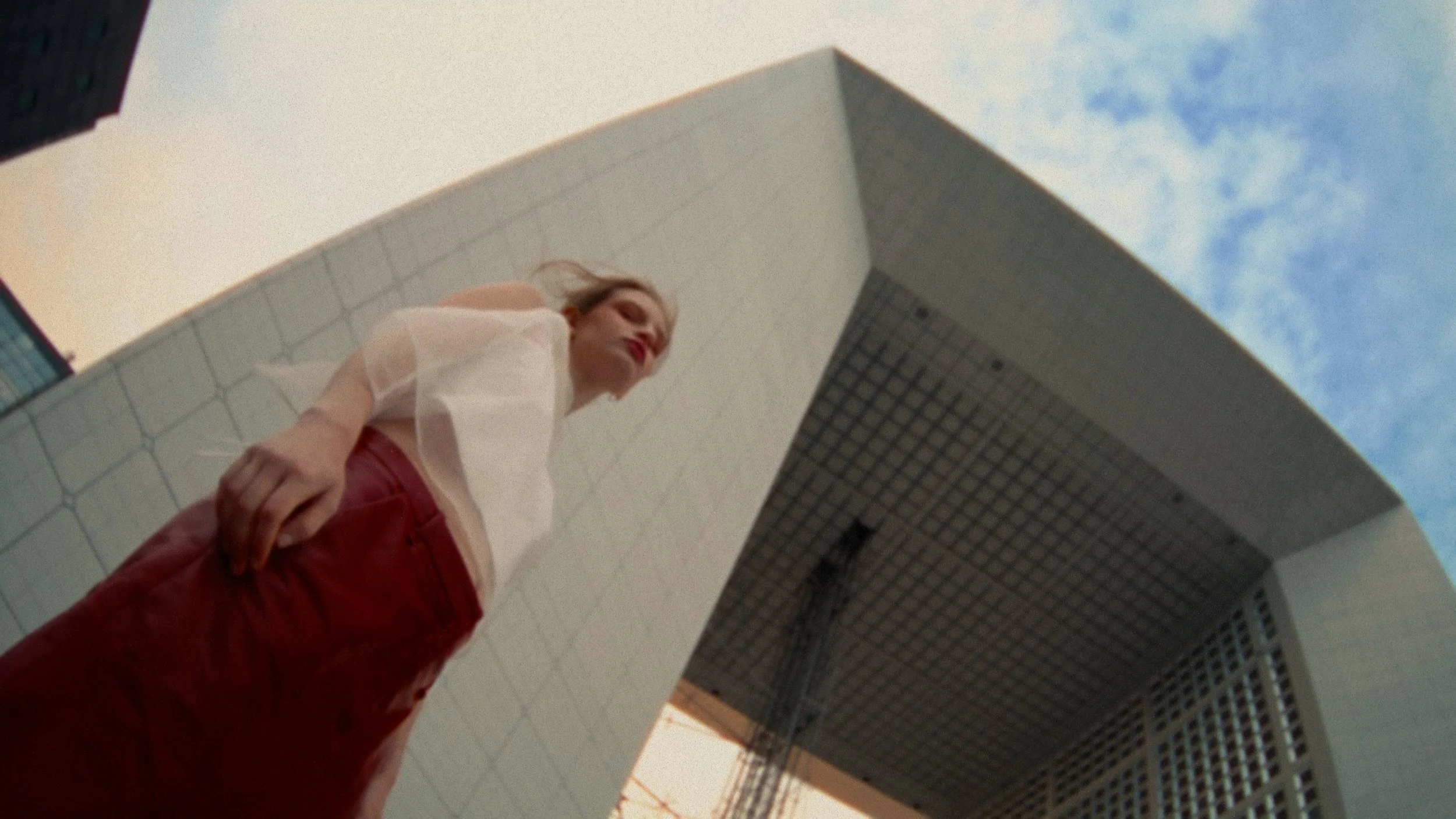 A woman with blonde hair and red lipstick standing under a modern, white architectural structure with a grid pattern ceiling, looking down at the camera, with a partly cloudy sky in the background.
