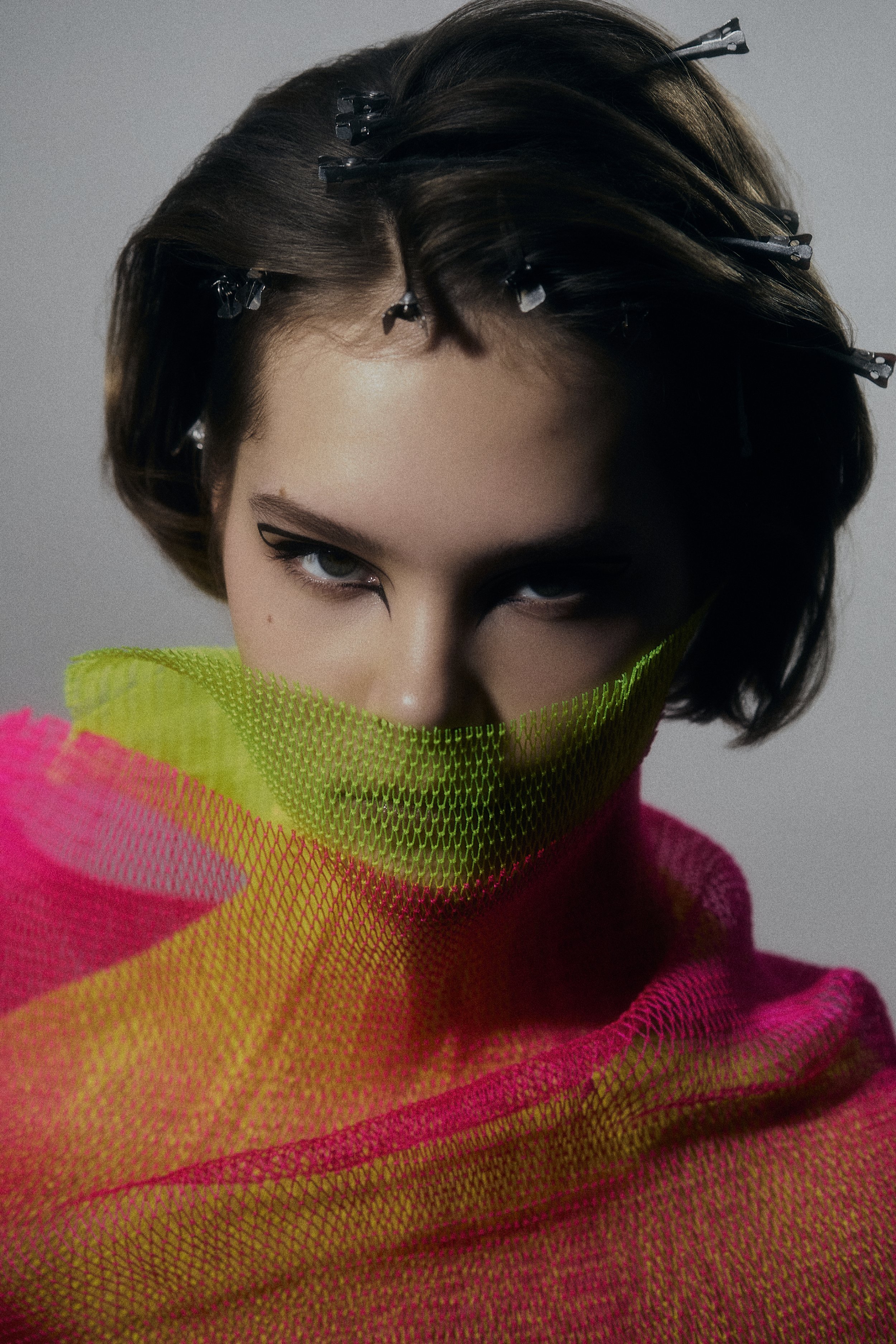 A woman with short, dark hair styled with hair clips, wearing dark makeup with winged eyeliner, a bright pink garment, and a neon yellow-green veil covering her mouth and part of her face.