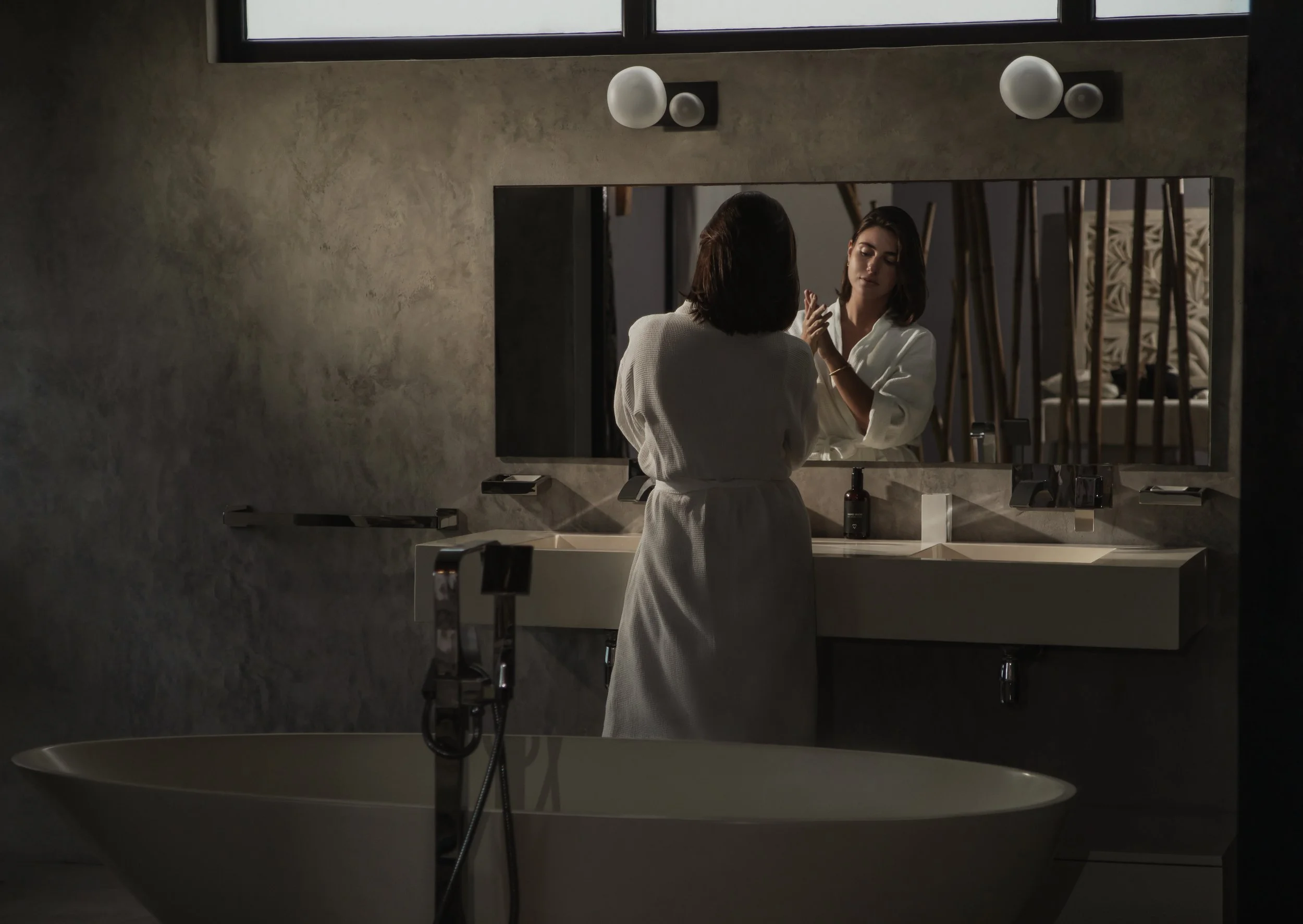 Woman in white robe applying makeup to her face in front of a mirror in a modern bathroom.