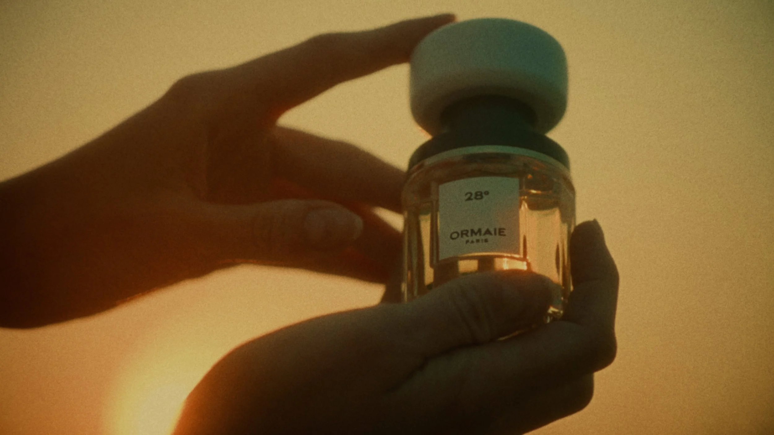 A person holding a bottle of Ormaie Paris perfume with a white cap against a plain background.