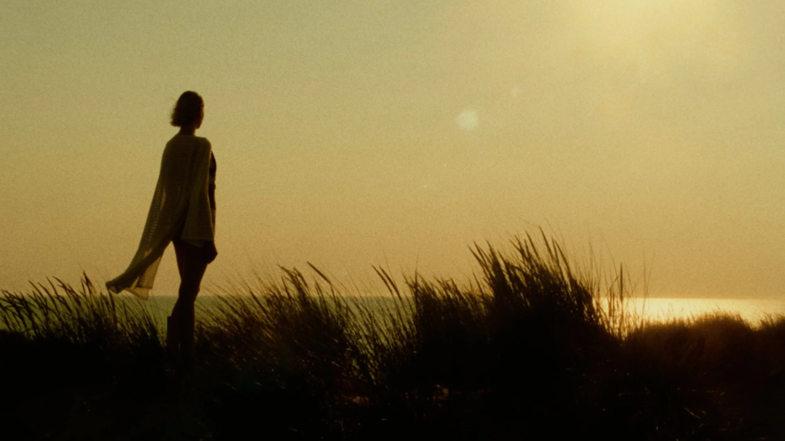 Silhouette of a woman walking through tall grass on a hill during sunset or sunrise, with a bright sky and the sun near the horizon.