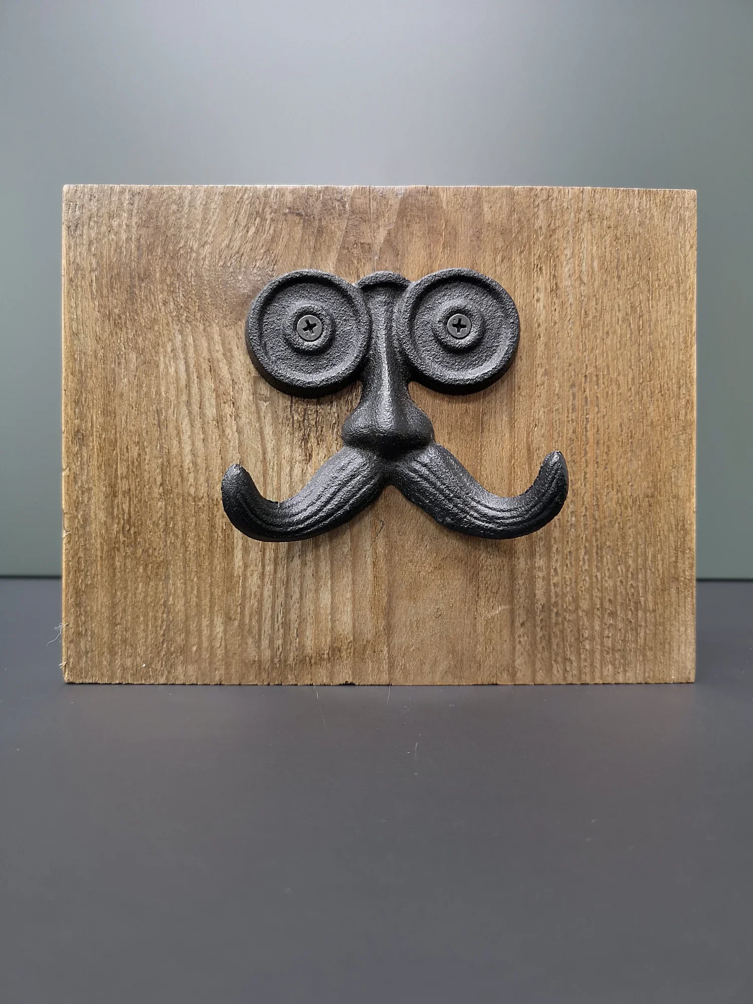 A wooden block with a black metal decorative piece resembling a face with large concentric circle eyes, a nose, and a mustache, attached with screws.