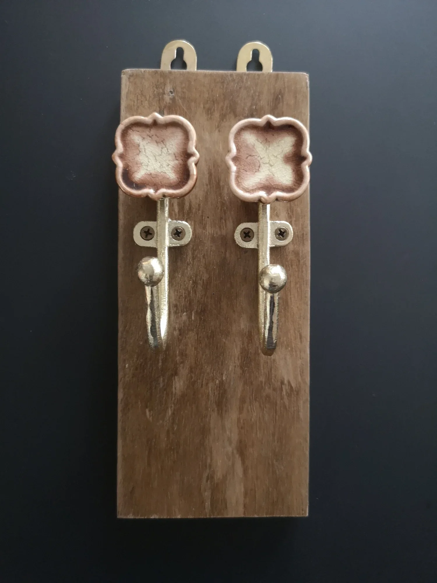 A pair of decorative earrings with a square-shaped, crackled pinkish-beige ceramic design, attached to metal hooks, mounted on a wooden display board.
