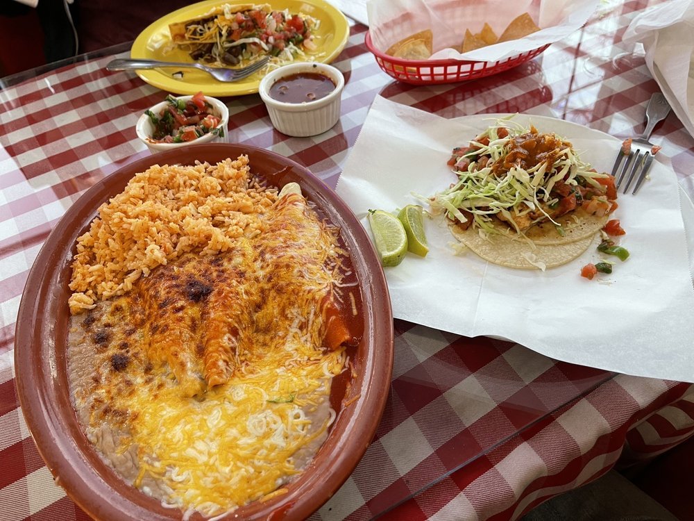 Mexican meal with a cheese and chicken enchilada casserole, Mexican rice, soft taco with shredded lettuce, tomatoes, and cheese, lime wedges, and sides of salsa and chips on a checkered red and white tablecloth.