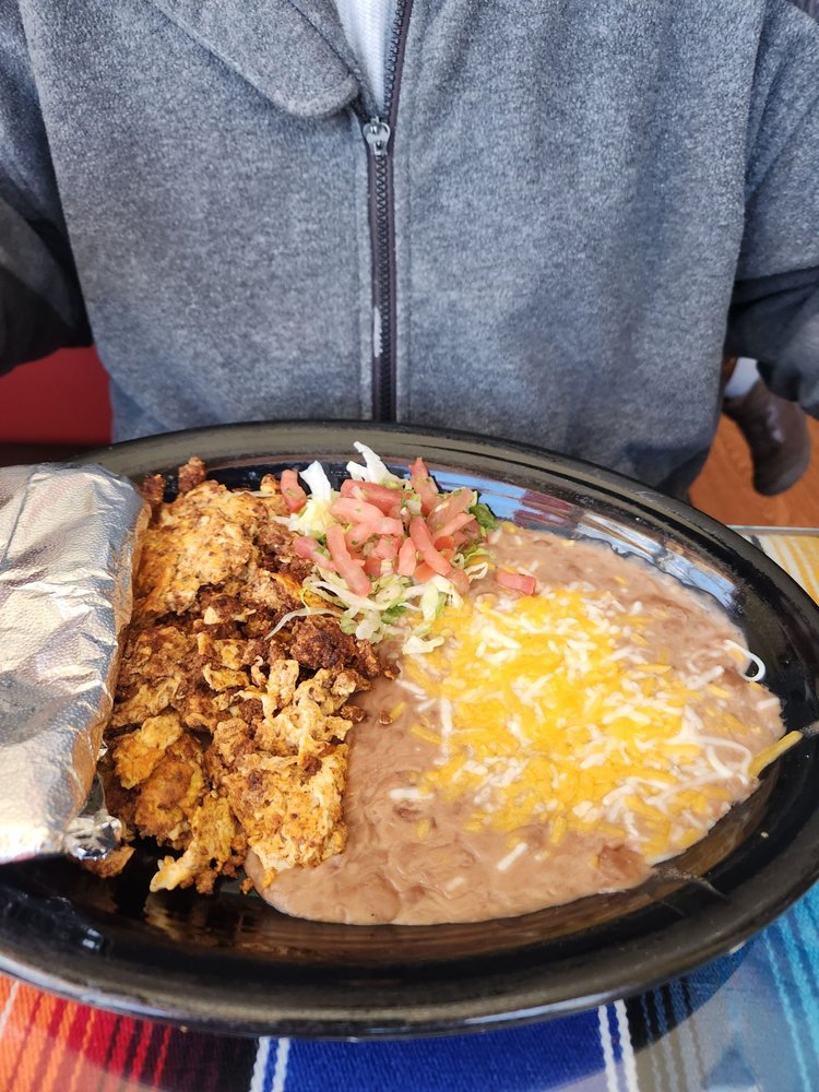 Plate of Mexican food with refried beans topped with shredded cheese, fried chicken, shredded lettuce, diced tomatoes, and chopped green onions, served with a foil-wrapped item on the side.