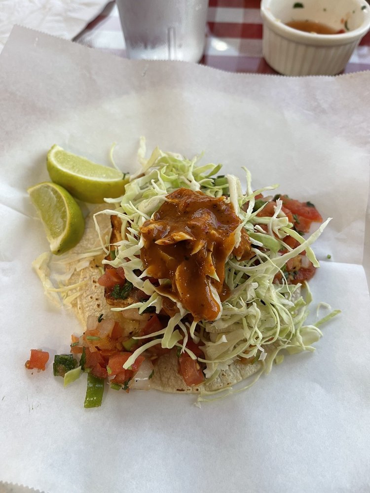Open fish taco topped with shredded cabbage, tomato salsa, and brown sauce, with lime wedges on the side, on white paper on a table.