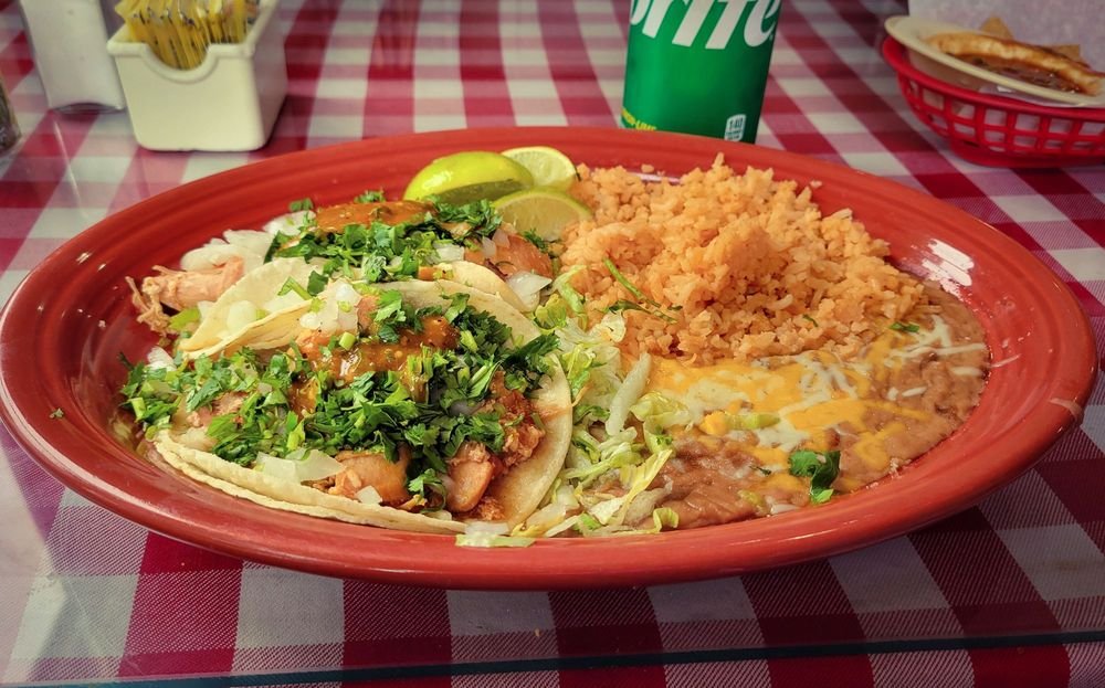 Plate of Mexican food with tacos, Mexican rice, refried beans with cheese, lime wedges, and a soda on a red checkered table.