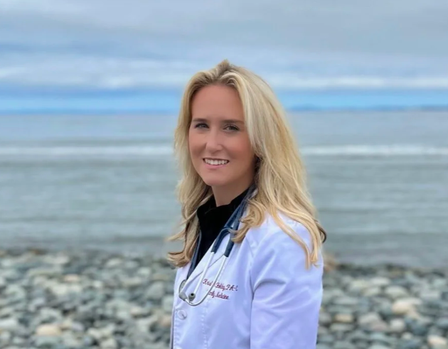 A blonde woman in a white medical coat with a stethoscope standing on a rocky beach with the ocean and cloudy sky in the background.