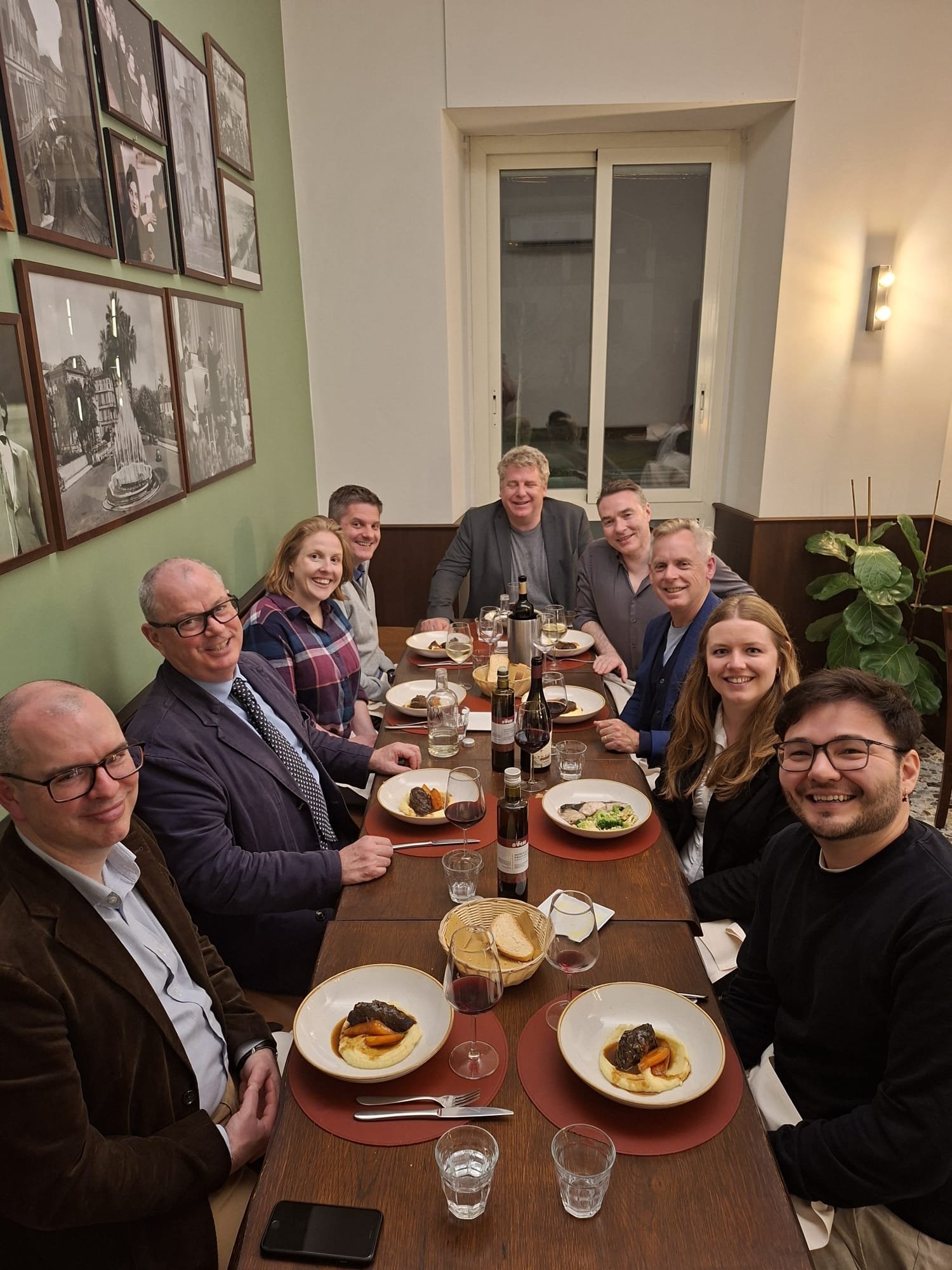 A group of ten people sitting around a dining table at a dinner party, smiling and enjoying their meal with wine and bread, in a cozy restaurant setting.