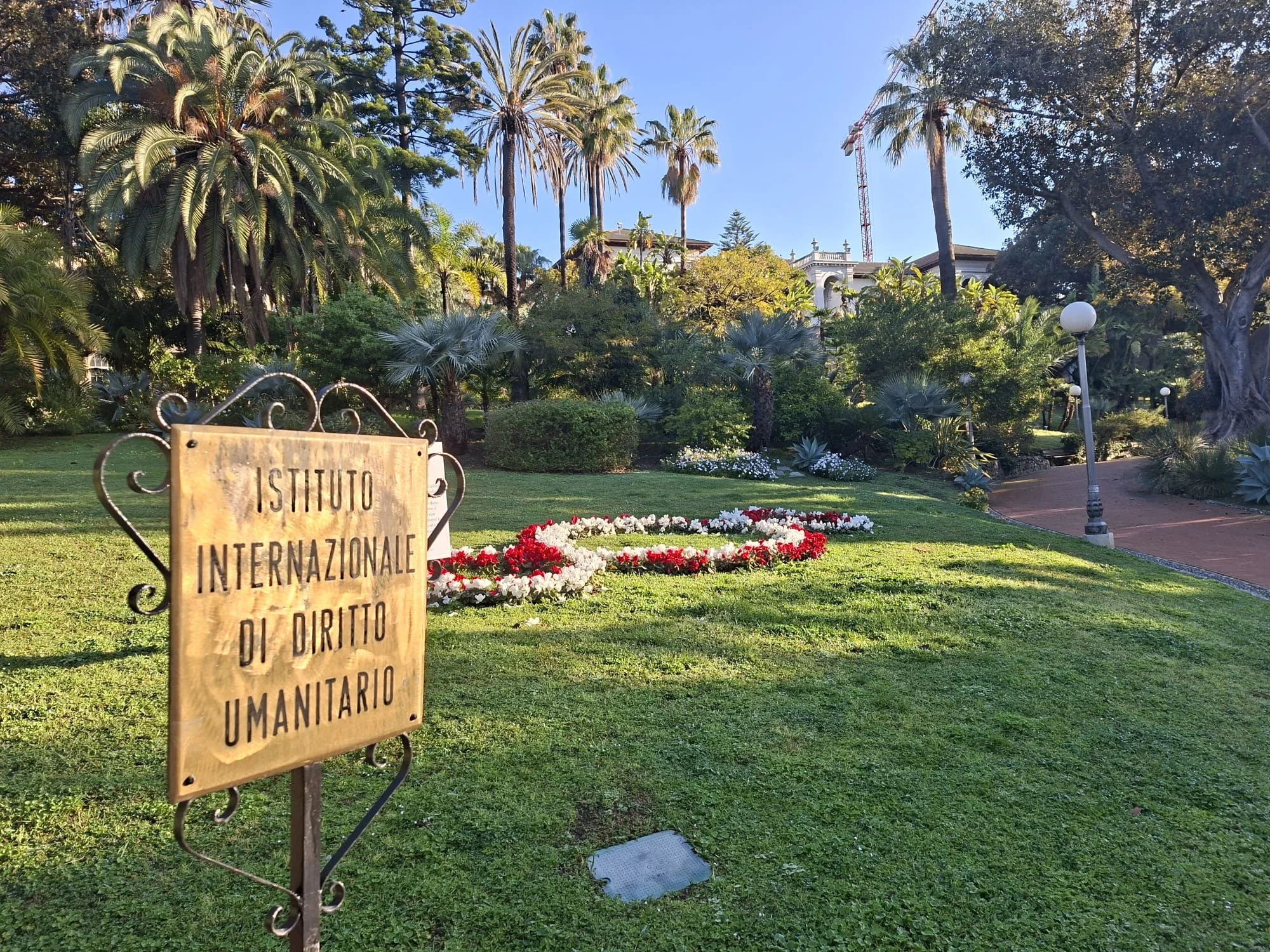 A garden with lush green grass, tall palm trees, and various plants and flowers. There is a wooden sign with Italian text, and the garden has a walkway with street lamps along it.