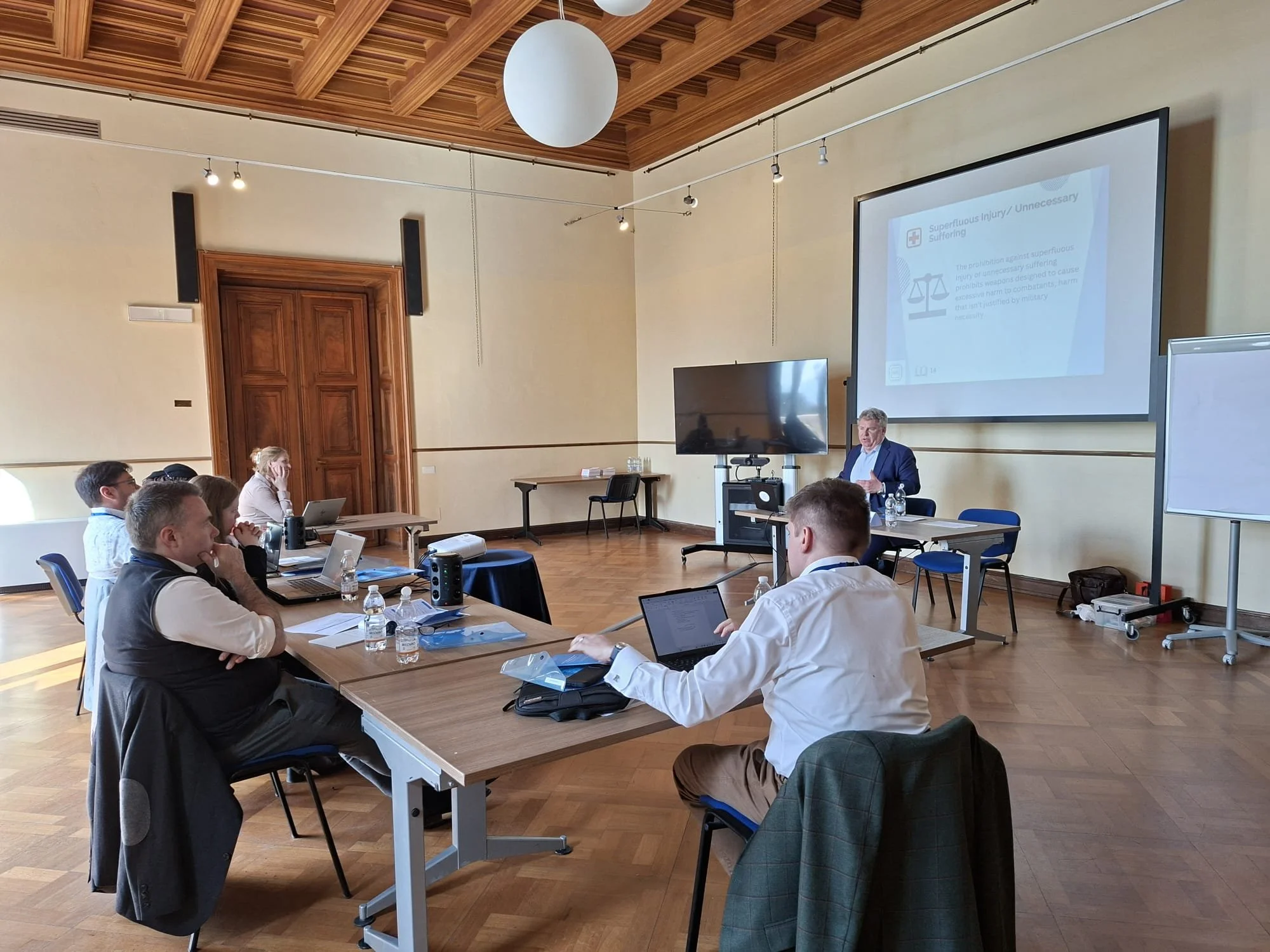 A group of people attending a business presentation or seminar in a conference room with wooden flooring and a decorative ceiling. The presenter is at the front, near a large screen displaying a PowerPoint slide. Attendees are seated at tables with l