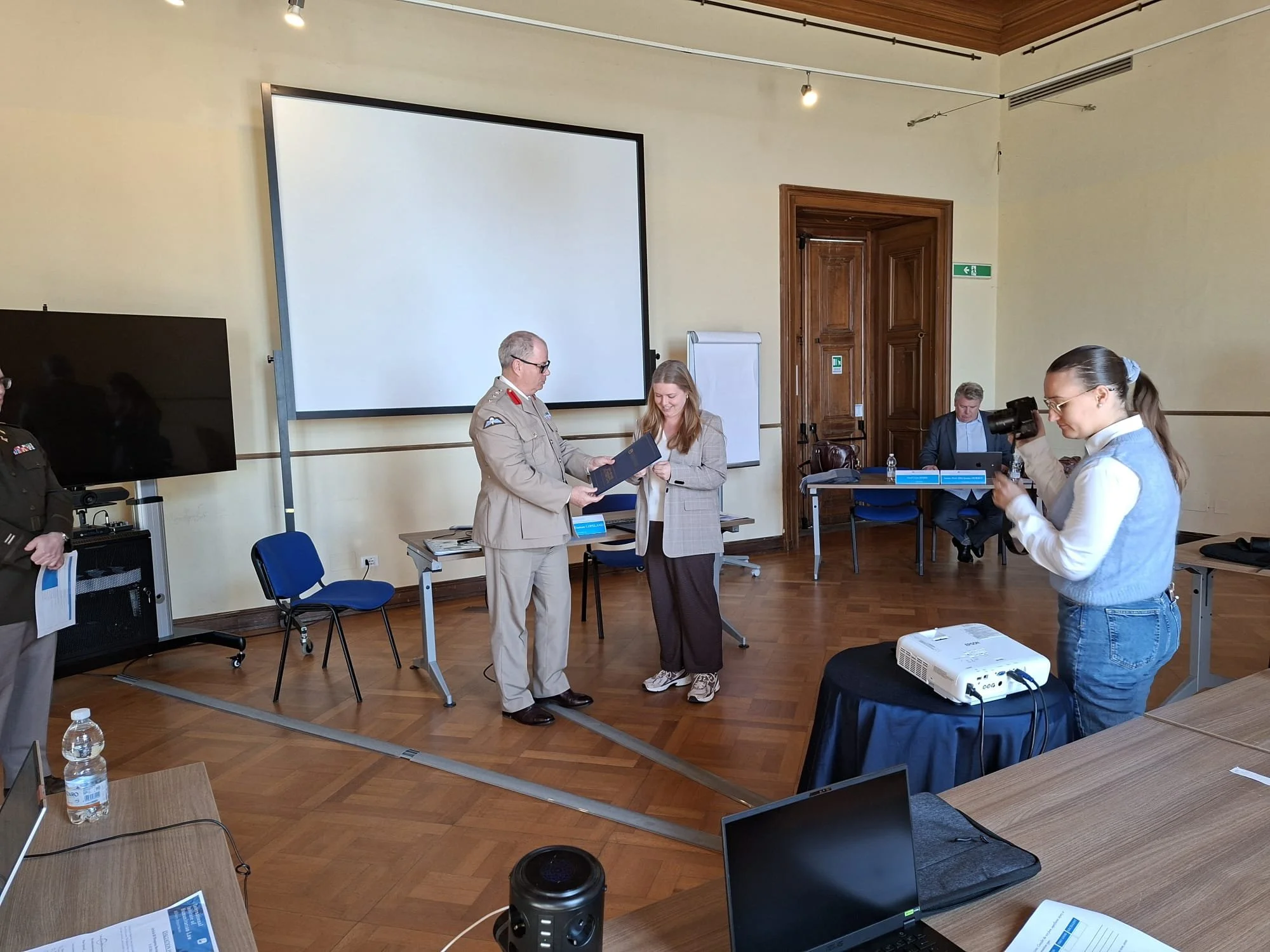 A woman is taking a picture of a man in a military uniform while a woman receives an award or certificate in a room with a large screen and a whiteboard.