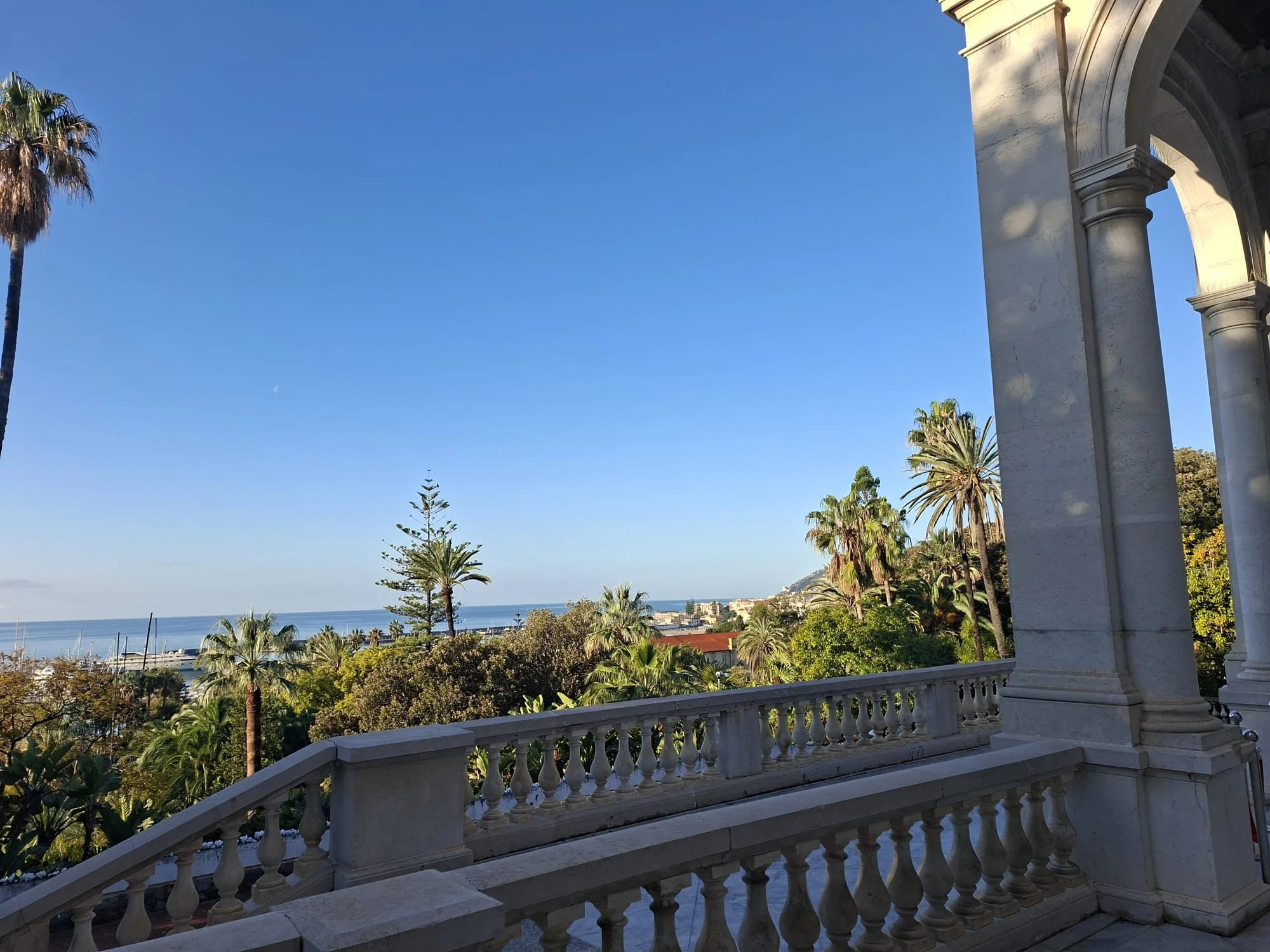 View from a stone porch with a white balustrade, overlooking a lush garden with palm trees, and a distant view of the ocean under a clear blue sky.