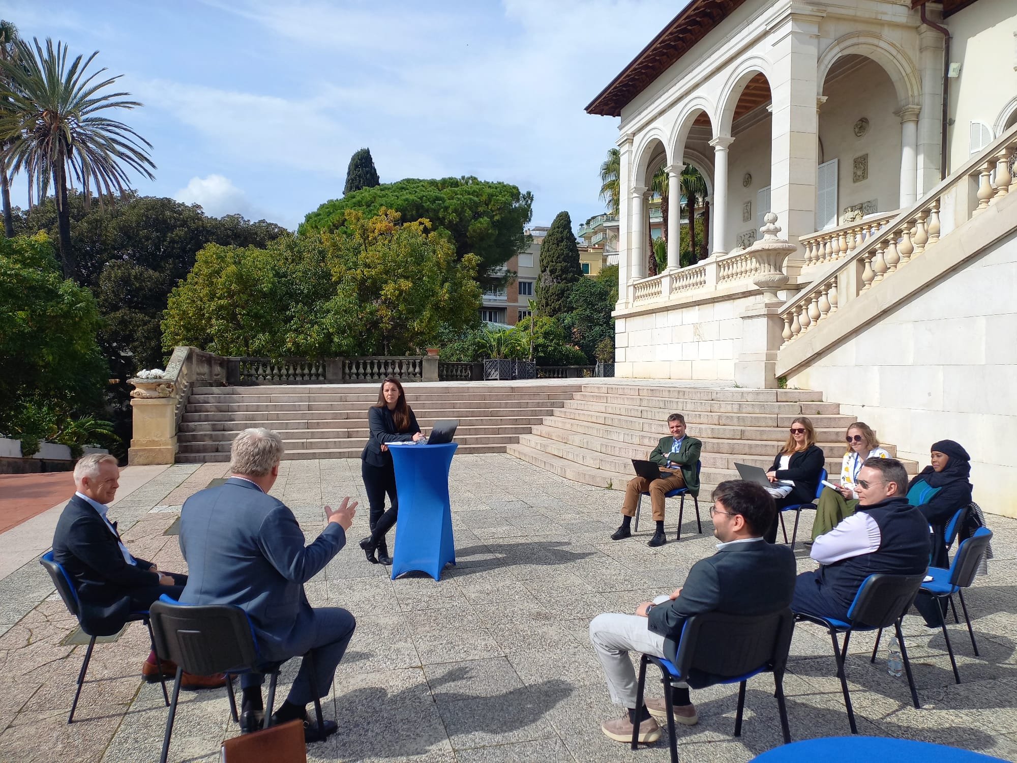 Outdoor business meeting or seminar with a woman presenting at a table, surrounded by men and women seated on chairs, in front of a grand white building with arches and stairs, lush green trees, and a blue sky.