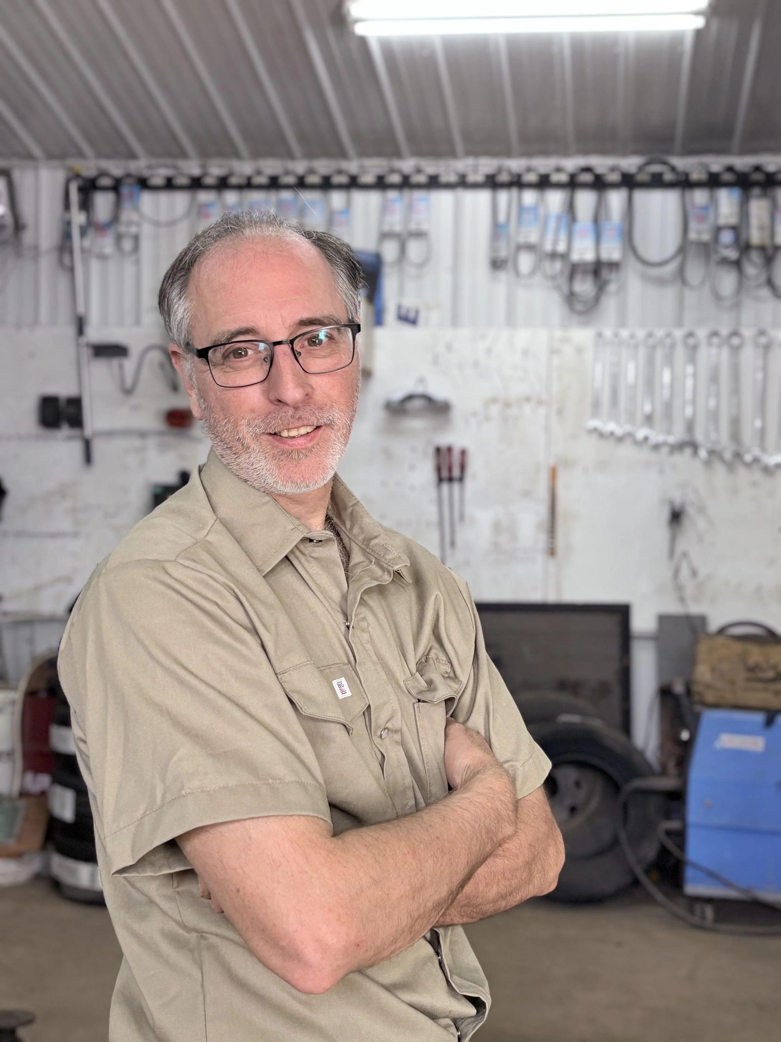 Un homme souriant avec des lunettes, portant une chemise beige à manches courtes, dans un atelier avec des outils accrochés au mur