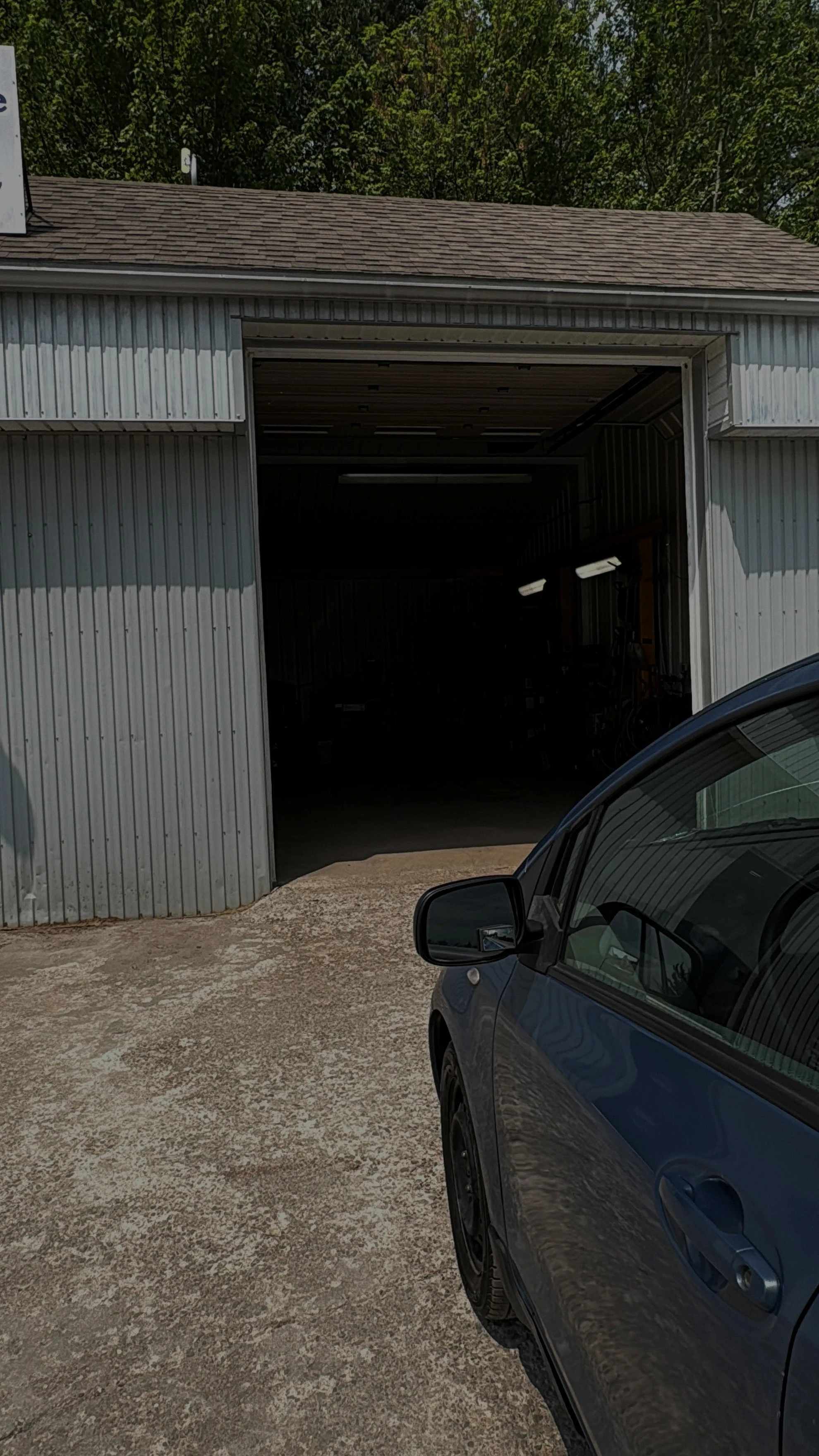 Une partie de voiture bleue devant un garage en métal, avec un sol en béton et des arbres en arrière-plan.