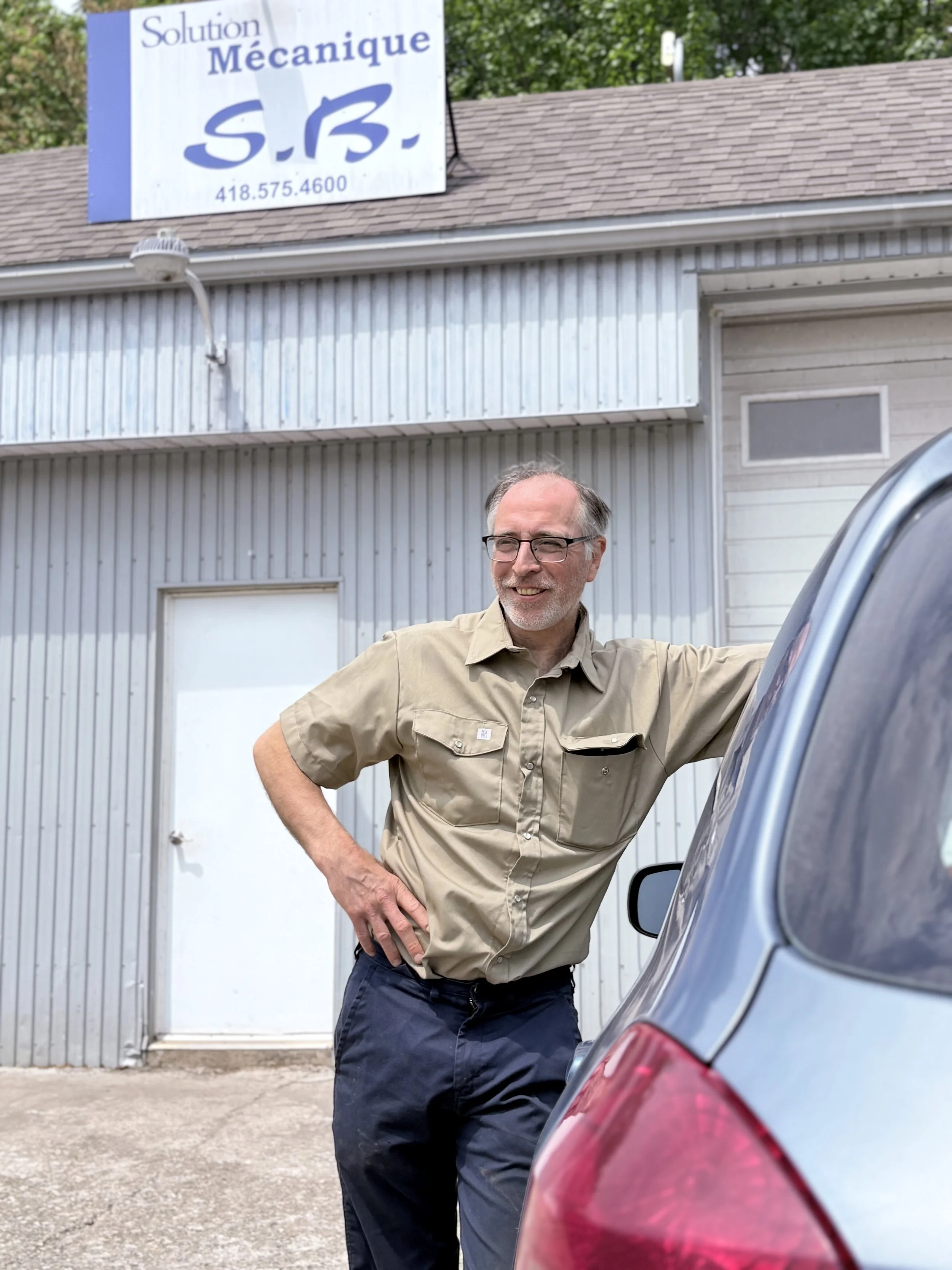 Un homme souriant portant une chemise beige et des lunettes, se tenant à côté d'une voiture, devant un bâtiment en métal gris avec une enseigne indiquant 'Solution Mécanique S.B.' et un numéro de téléphone.