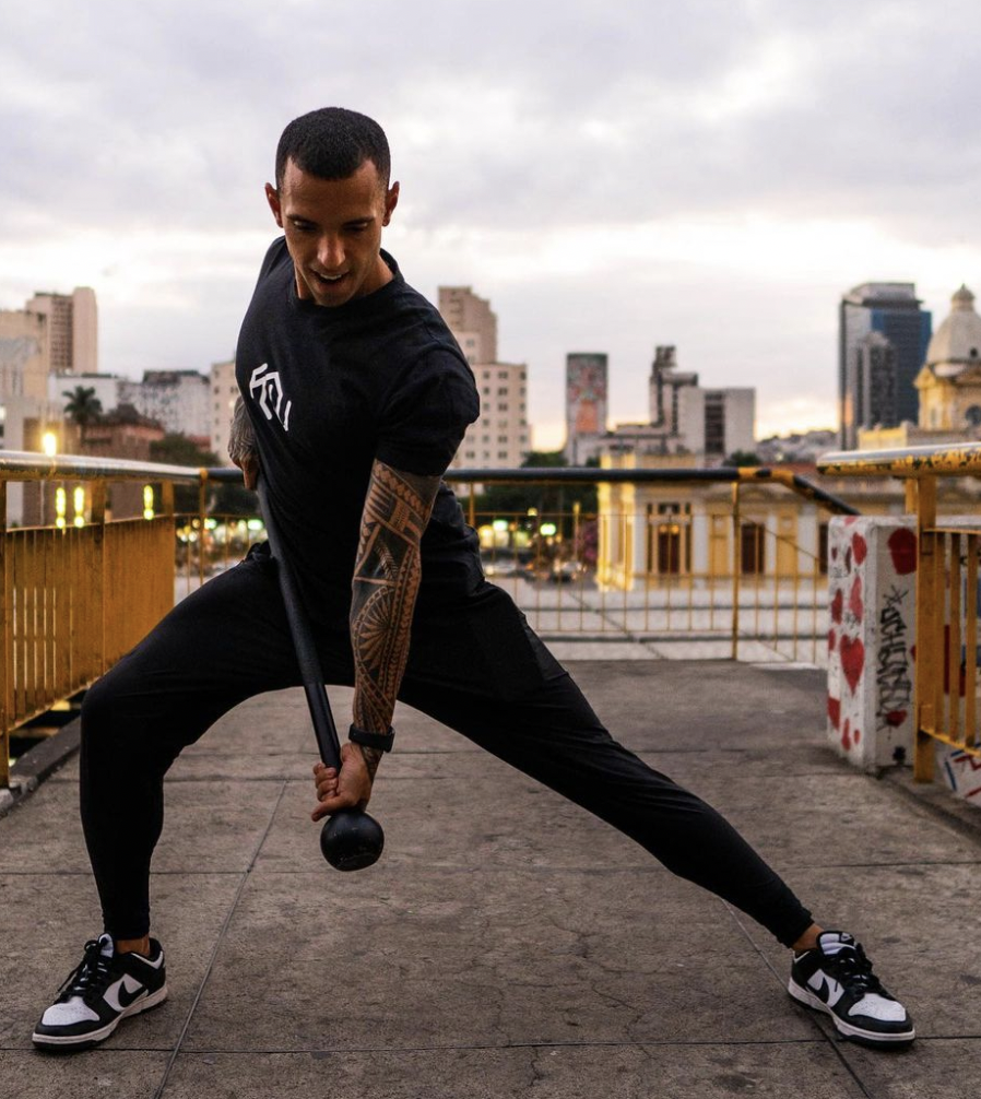 A man working out on an outdoor rooftop with an urban cityscape in the background at sunset, holding a mace weight with both hands and spreading his legs apart in a wide stance.